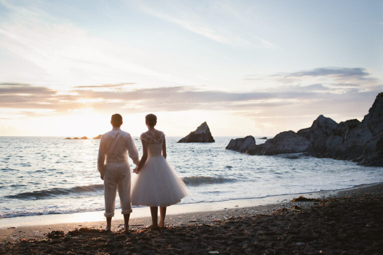 A bride and groom standing hand in hand looking across the sea at Tunnels Beaches on their wedding day