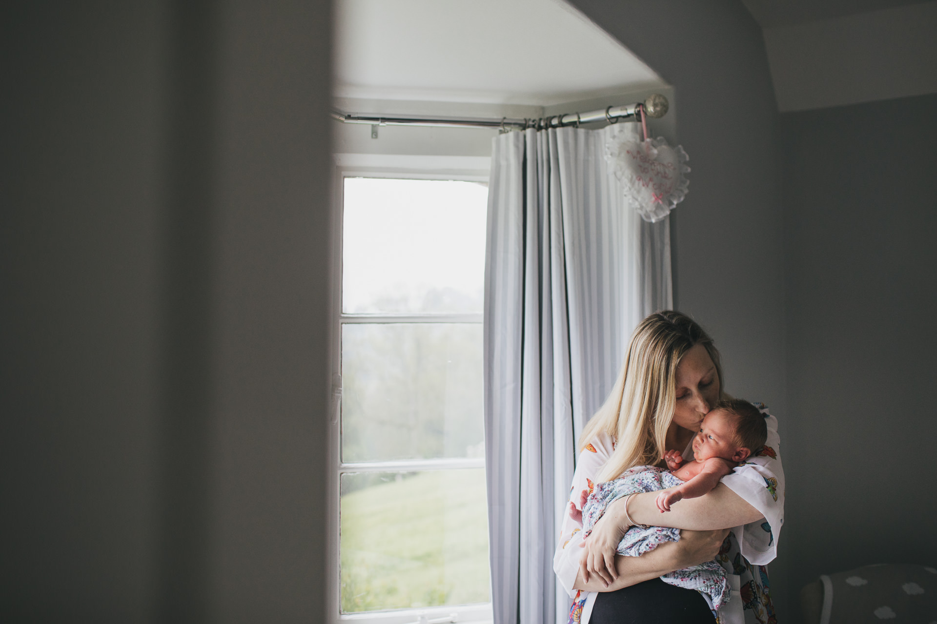 Mother cuddling newborn baby by a large window during a newborn photography session in Devon