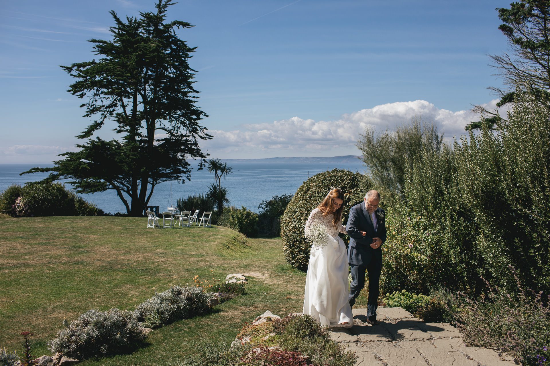 Bride and father walking across lawn with sea behind