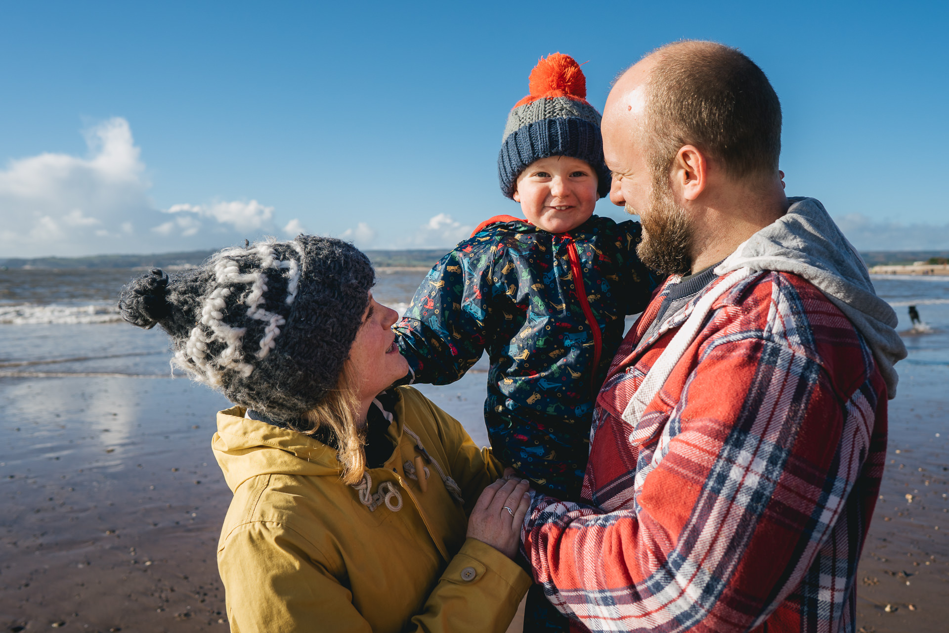 A young child in a bobble hat on a beach, held by his mum and dad
