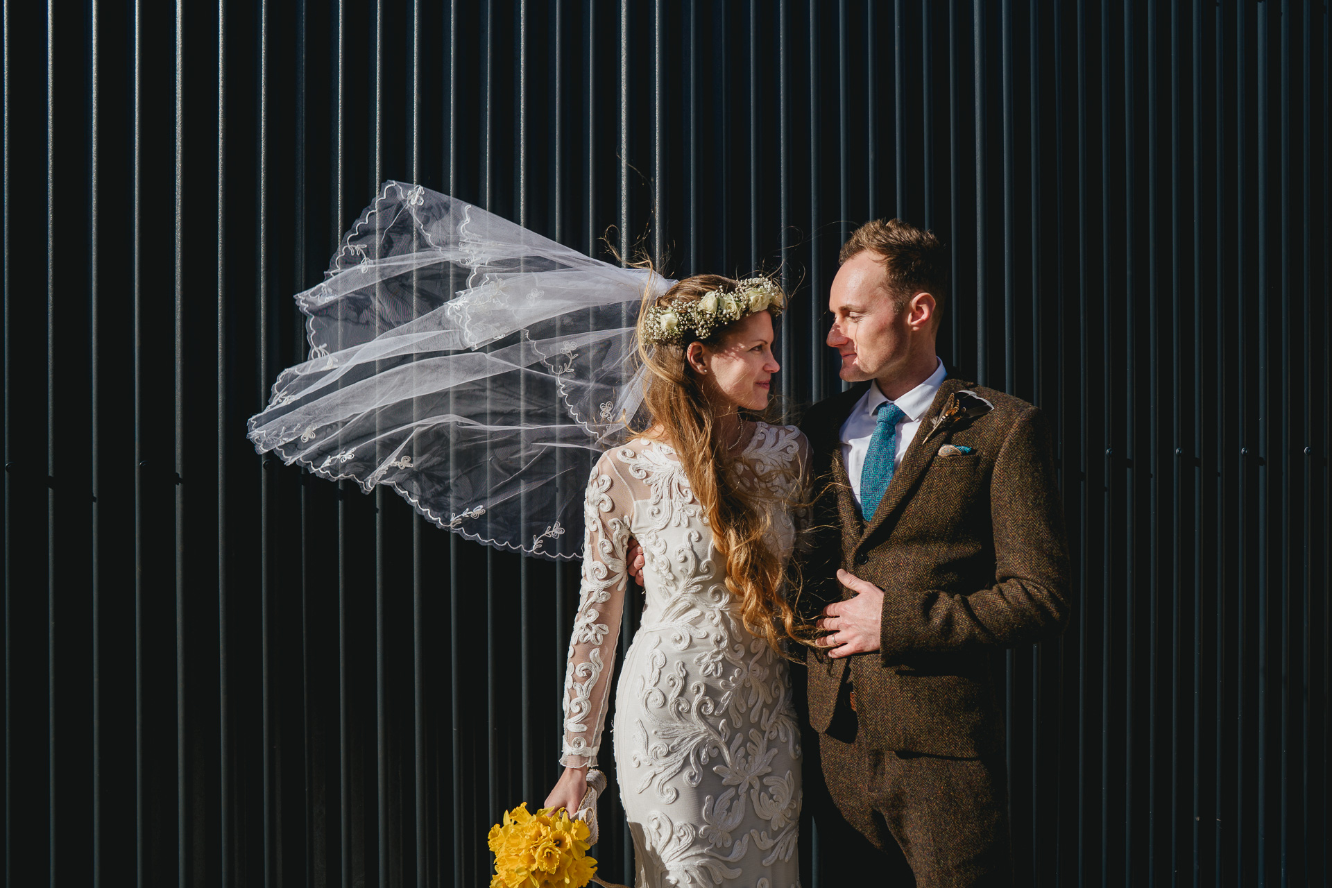 A bride and groom standing in front of a corrugated shed, with the bride's veil blowing in the wind