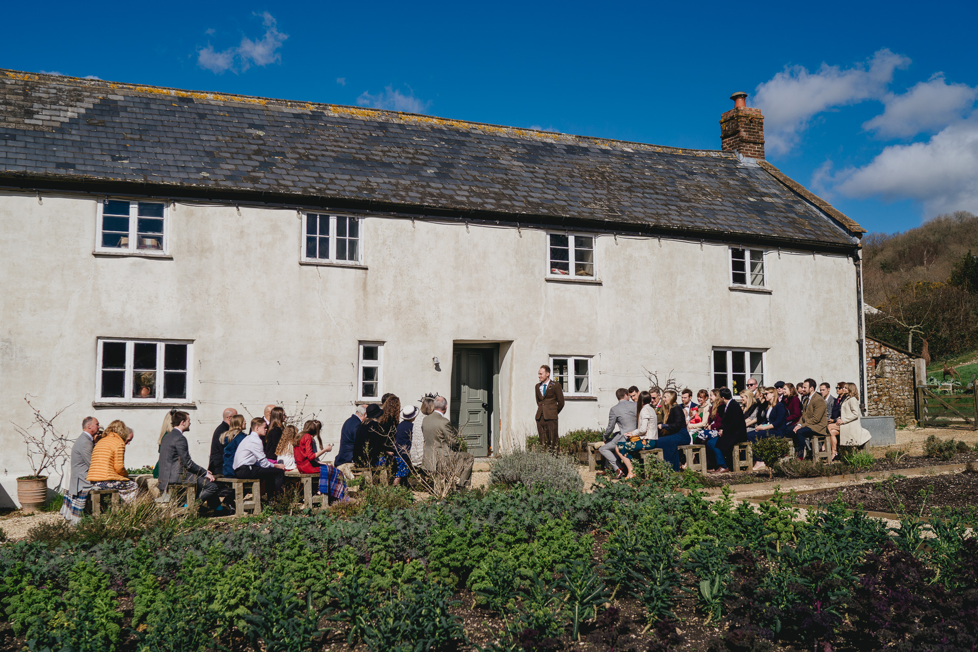 A groom standing outside River Cottage with wedding guests seated around him