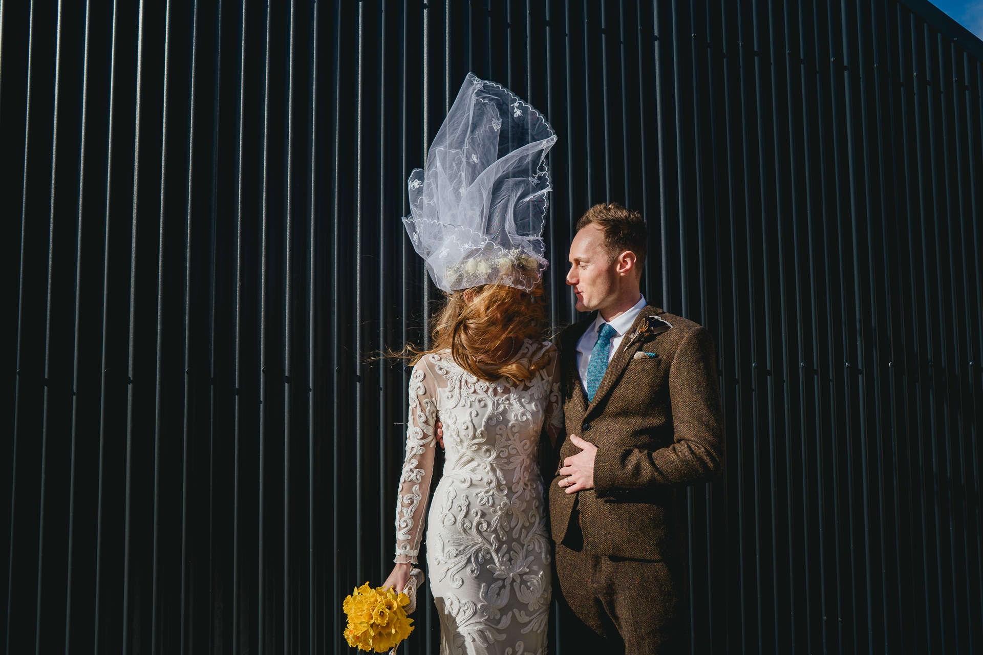 A bride and groom standing in front of a corrugated shed, with the bride's veil blowing in the wind
