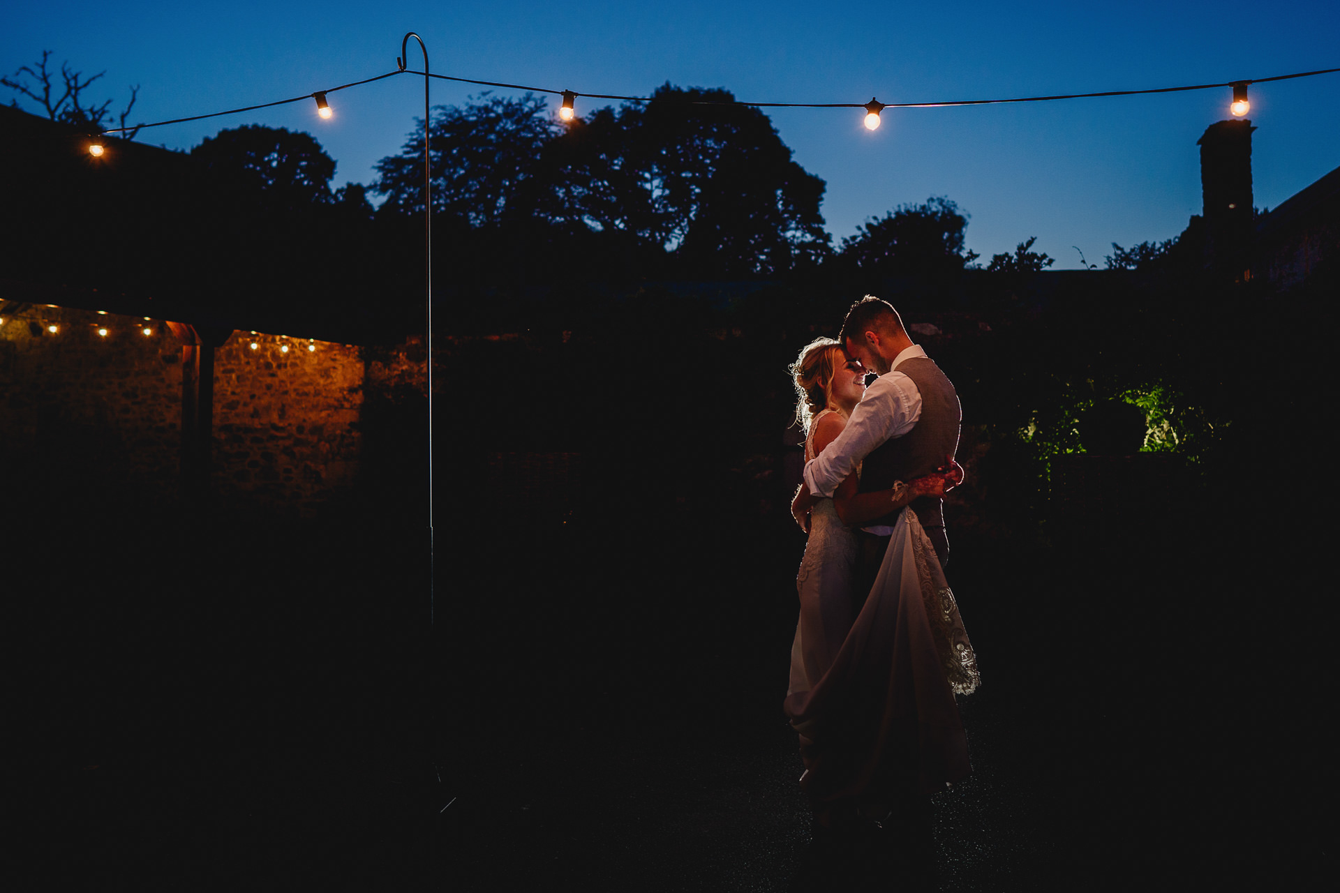 Bride and groom kissing under festoon lights at night