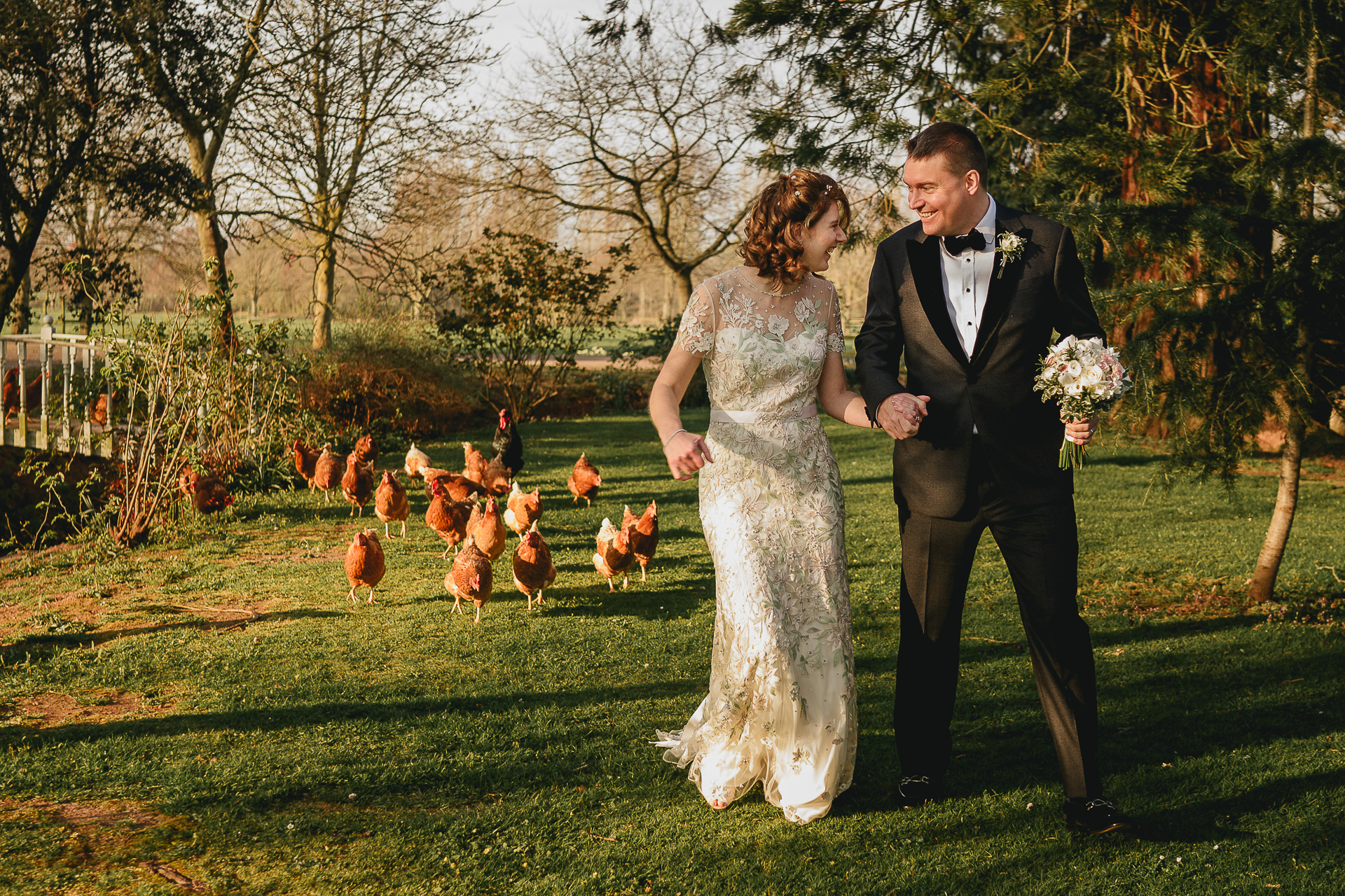 A bride and groom laughing with a group of chickens chasing them