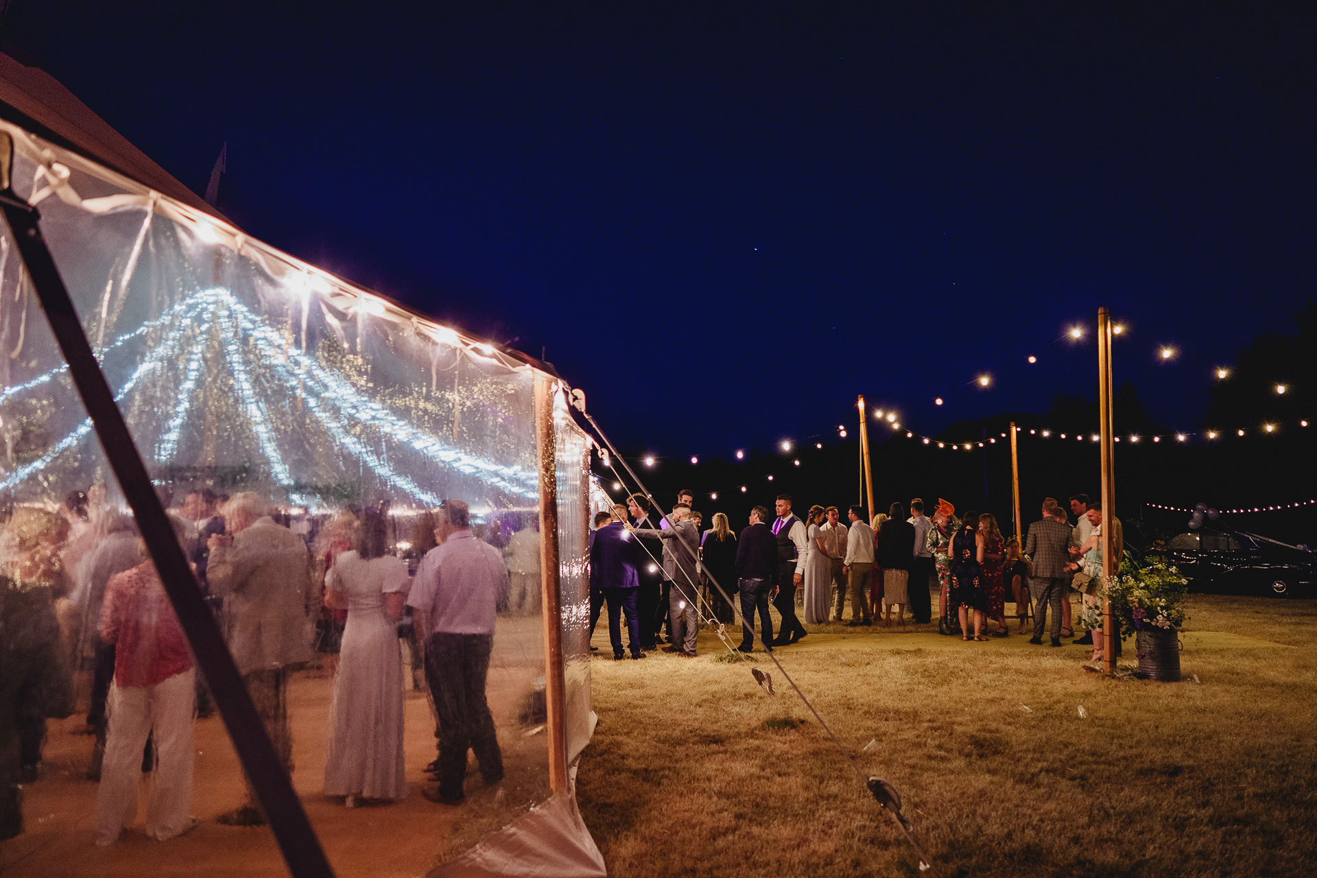 A marquee at night, with guests inside and out amongst festoon lighting