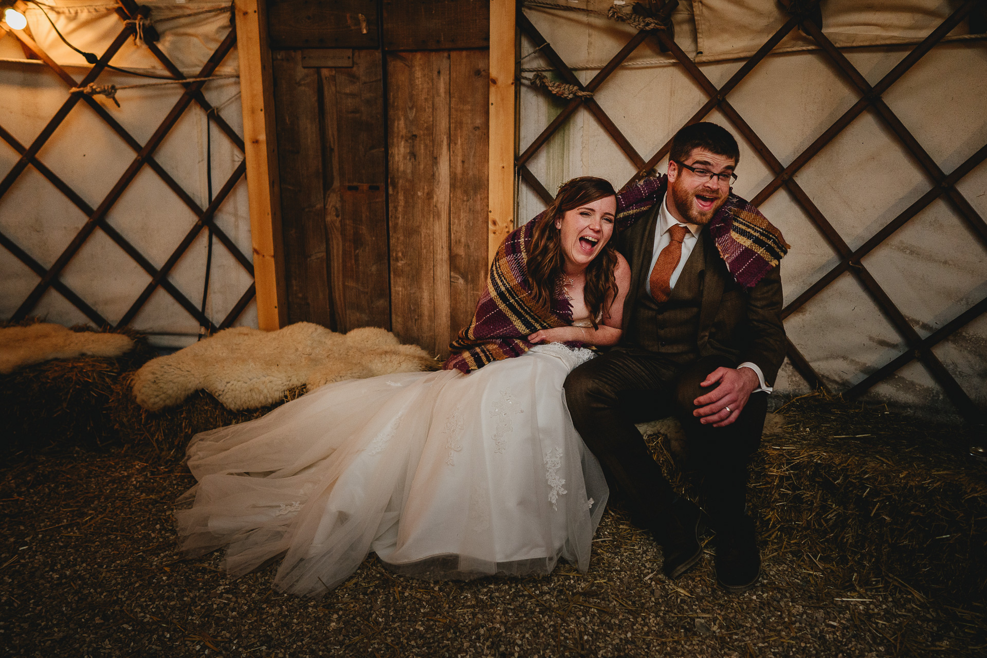 Bride and groom sitting on hay bales wrapped in a blanket laughing together