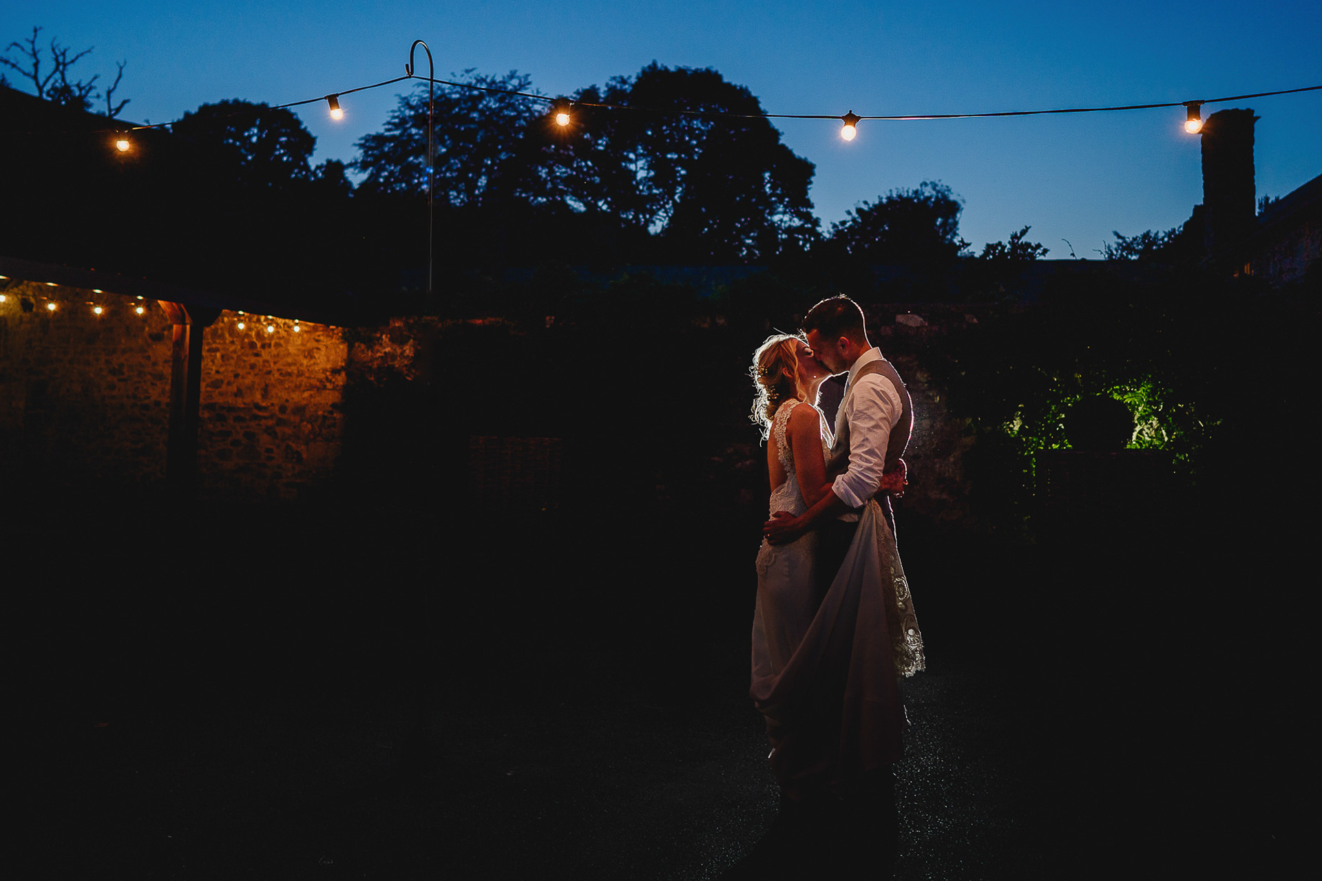 Bride and groom kissing underneath festoon lighting at night
