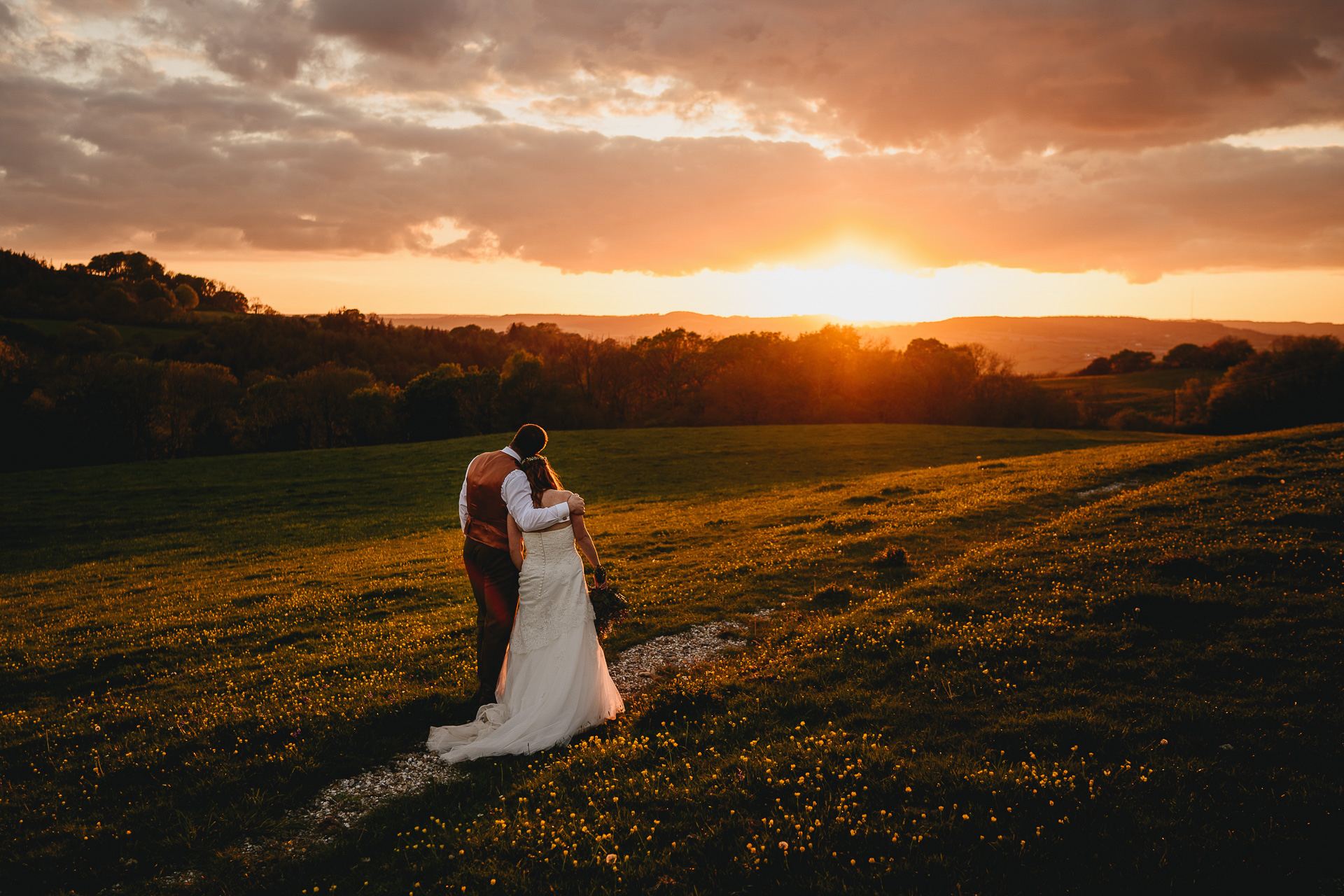 A bride and groom standing together and watching a sunset over countryside