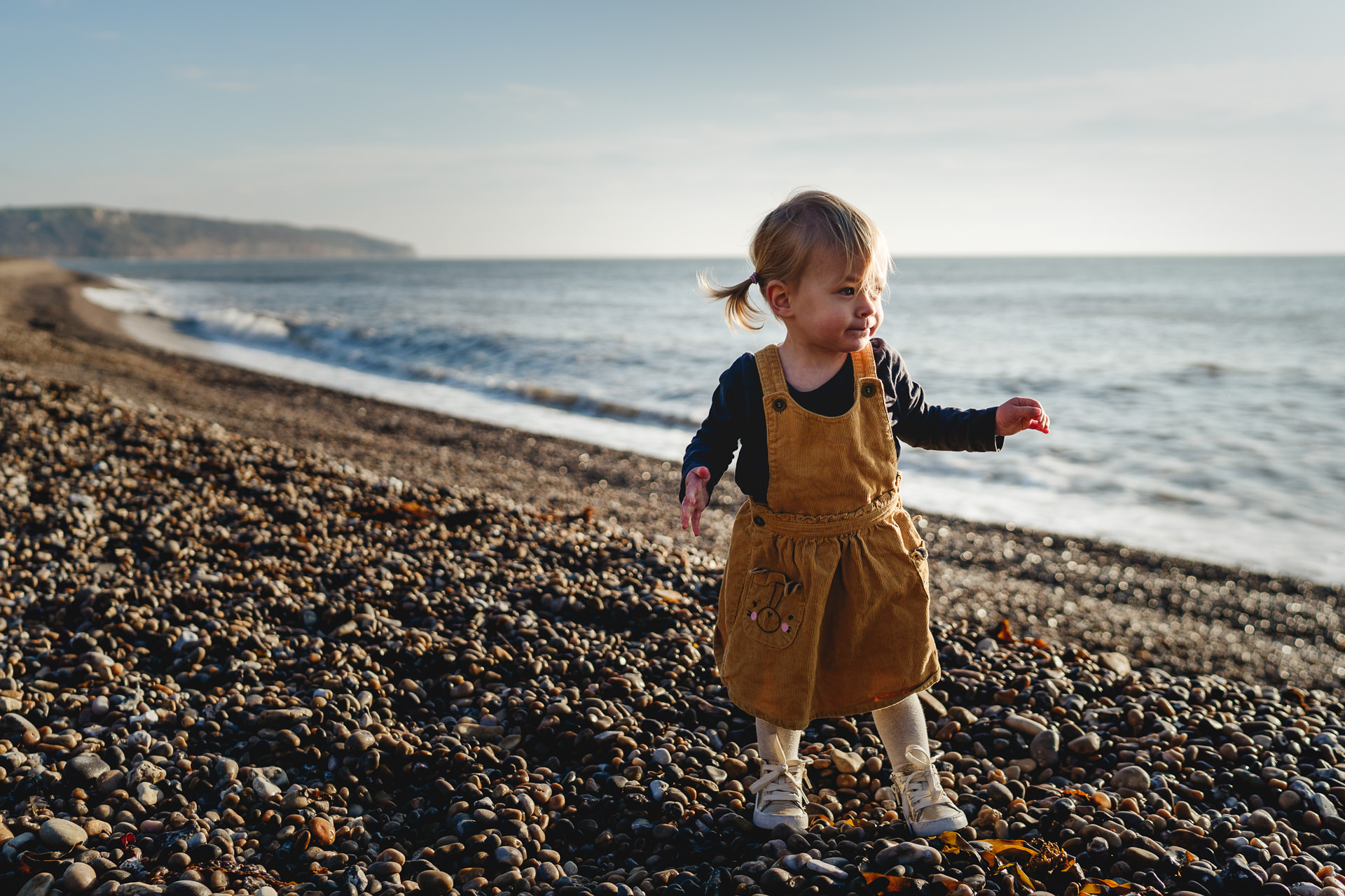 A toddler girl in a yellow dress on a pebble beach