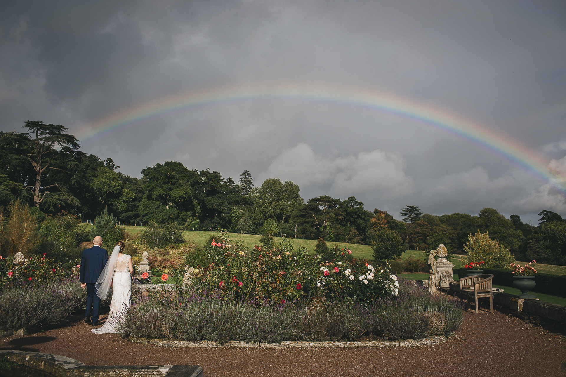 A bride and groom walking through a rose garden with a rainbow above them on a wet weather wedding day. 