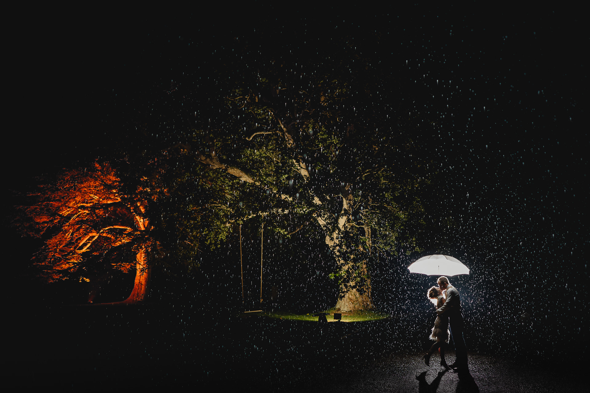 A rainy wedding day portrait, with a couple kissing in the dark, in heavy rain, with two large trees lit up behind them. 