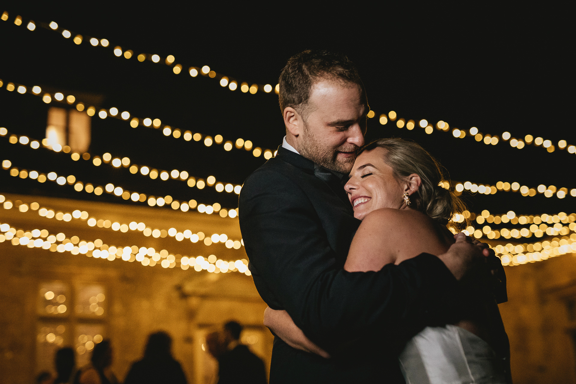 Bride and groom smiling and cuddling with festoon lights behind