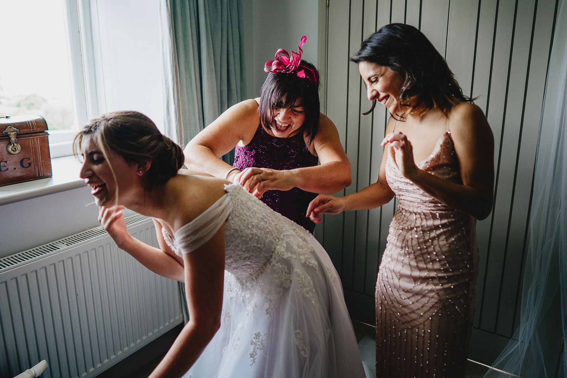 Mother and bridesmaid laughing whilst helping bride with dress