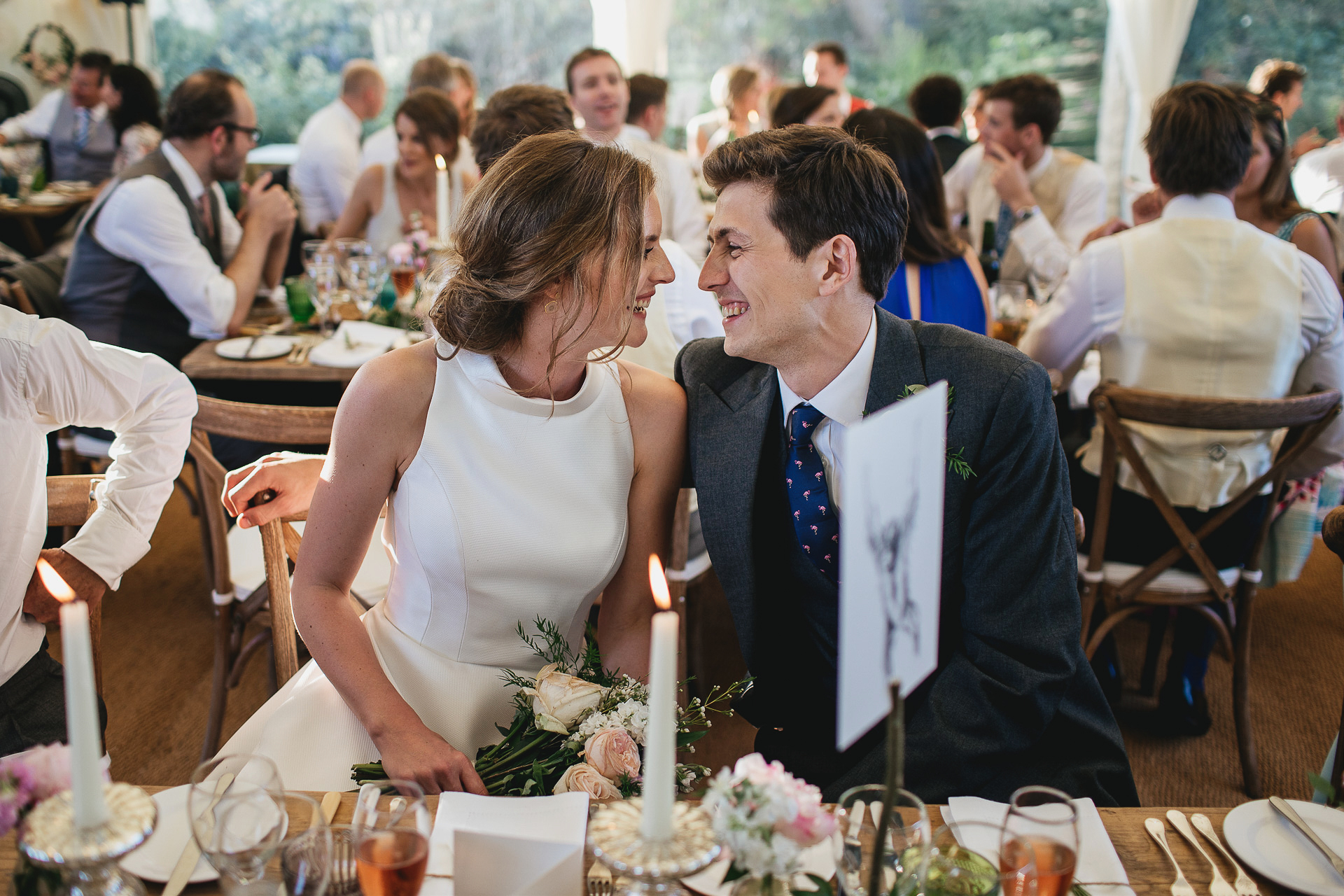 Bride and groom smiling at each other whilst seated in a marquee
