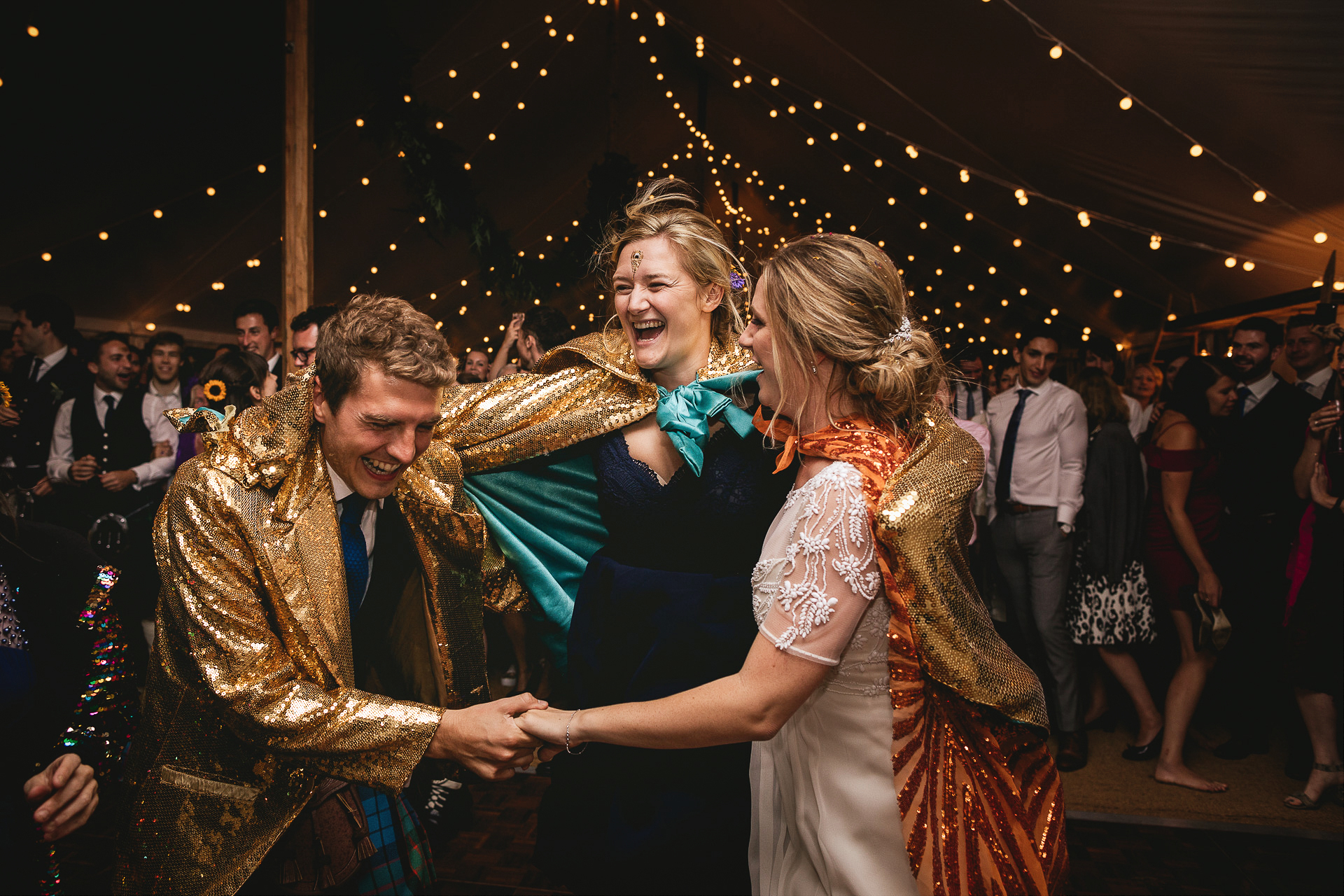 Bridesmaid leaping onto dance floor with bride and groom in sparkly capes