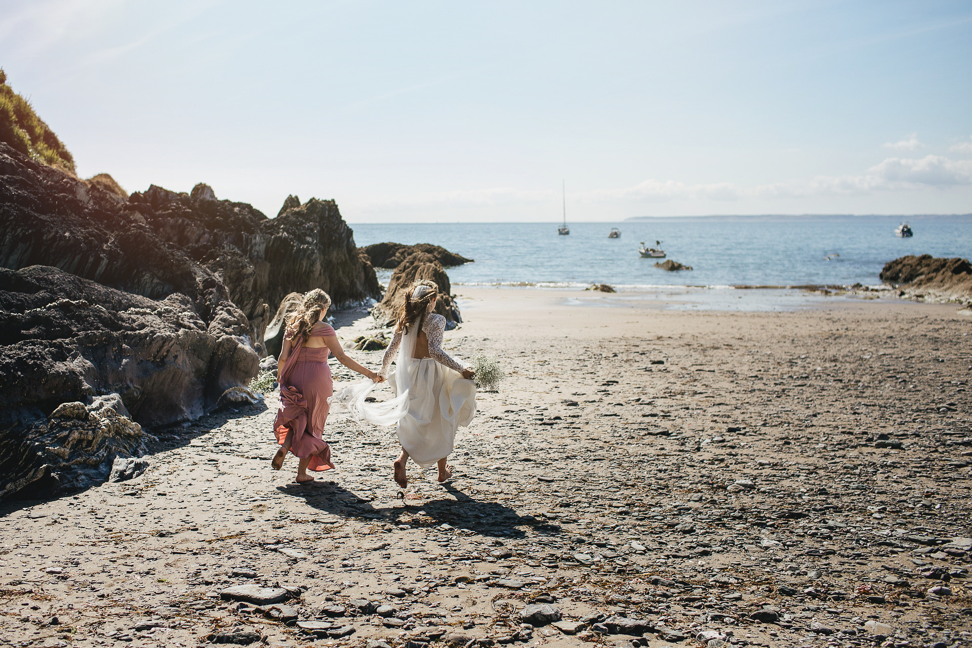 Bride and bridesmaid running hand in hand barefoot down the beach