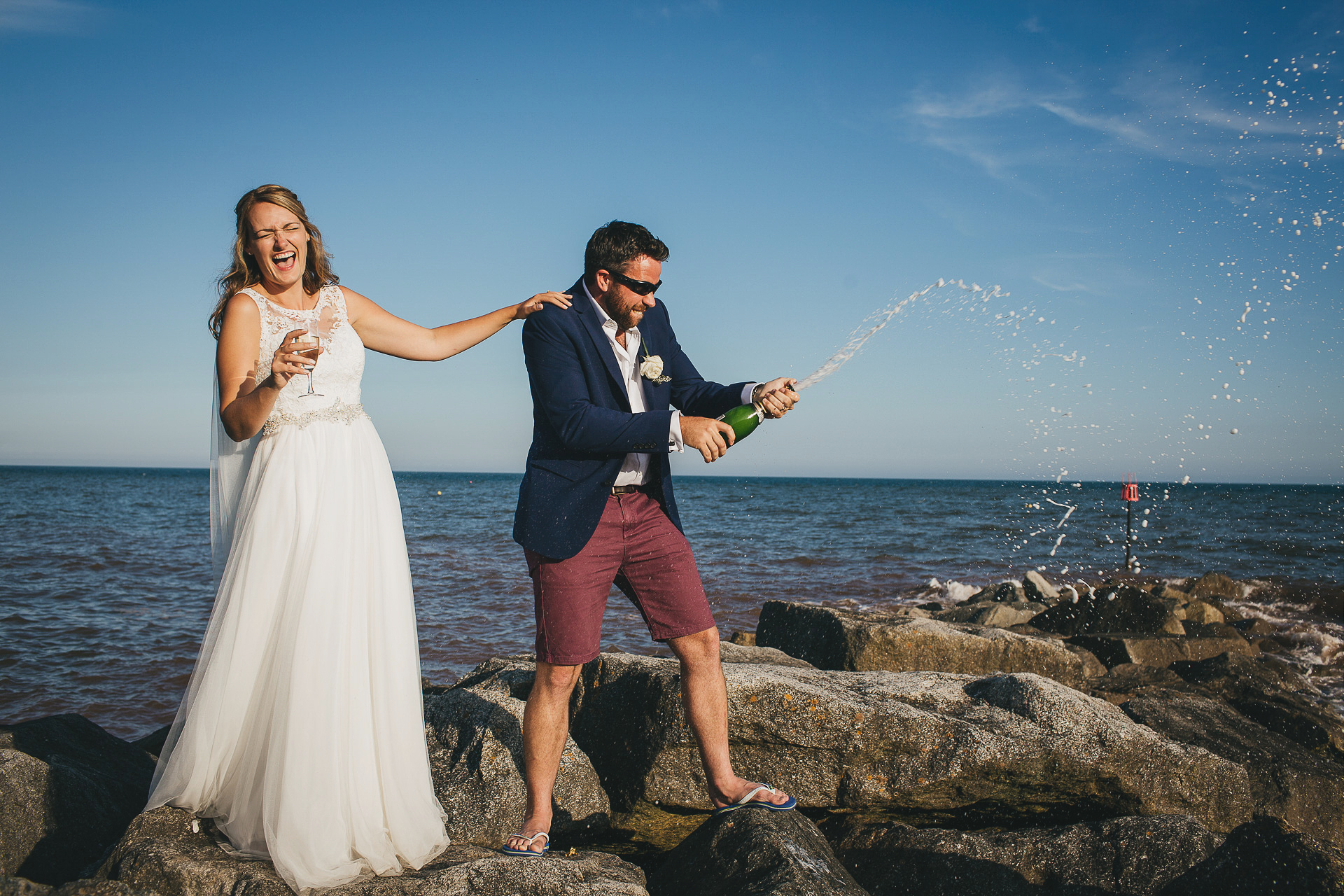 Bride and groom laughing and popping champagne on rocks by the sea