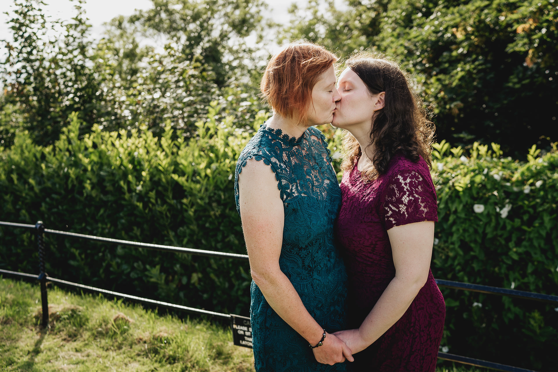 Two brides in colourful dresses kissing in sunshine