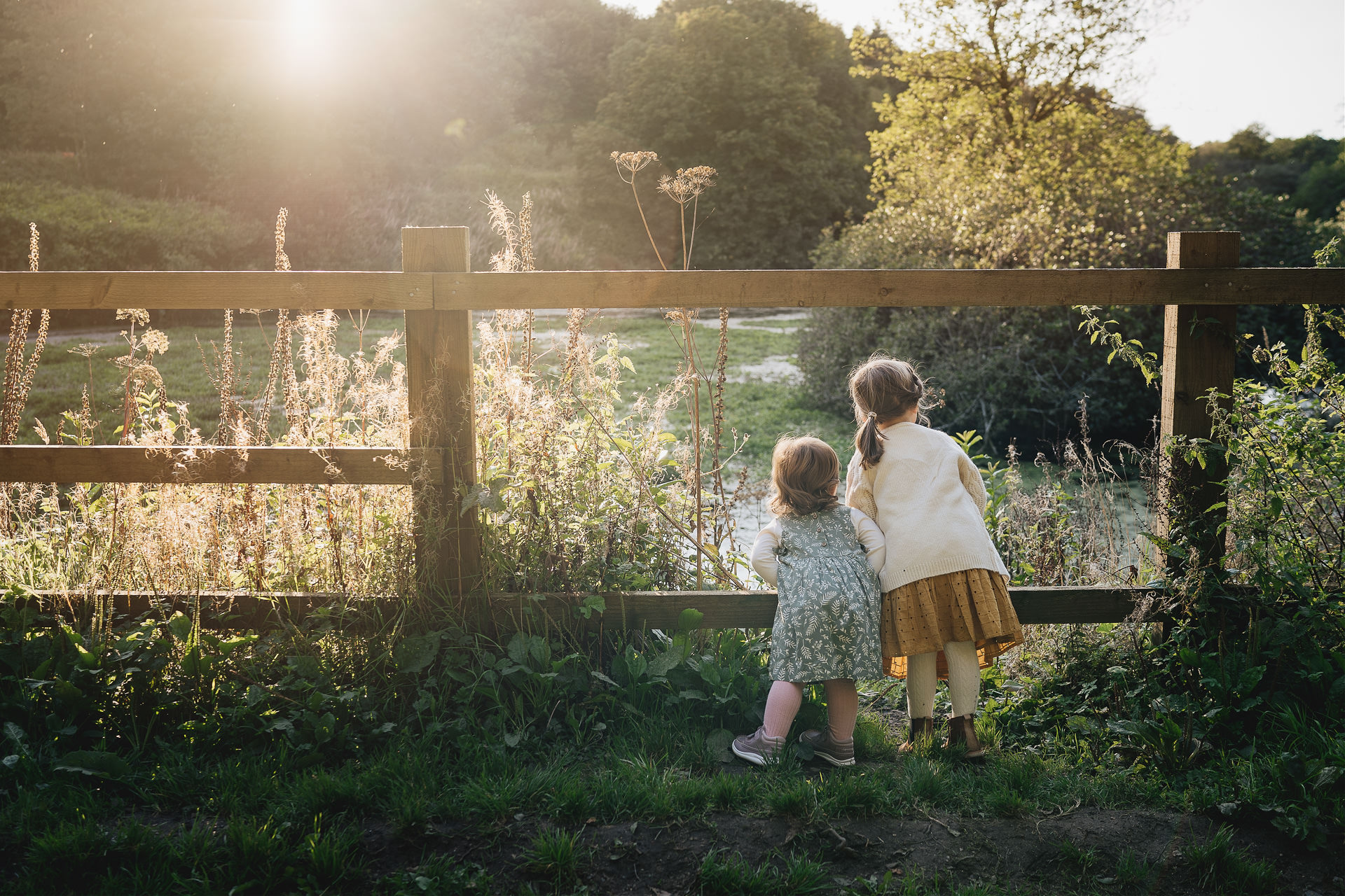 Two children peeping through a fence at a lake in evening sunlight
