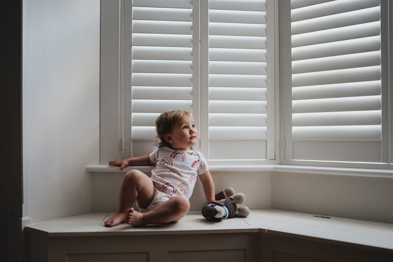 A toddler sitting on a window sill with wooden shutters