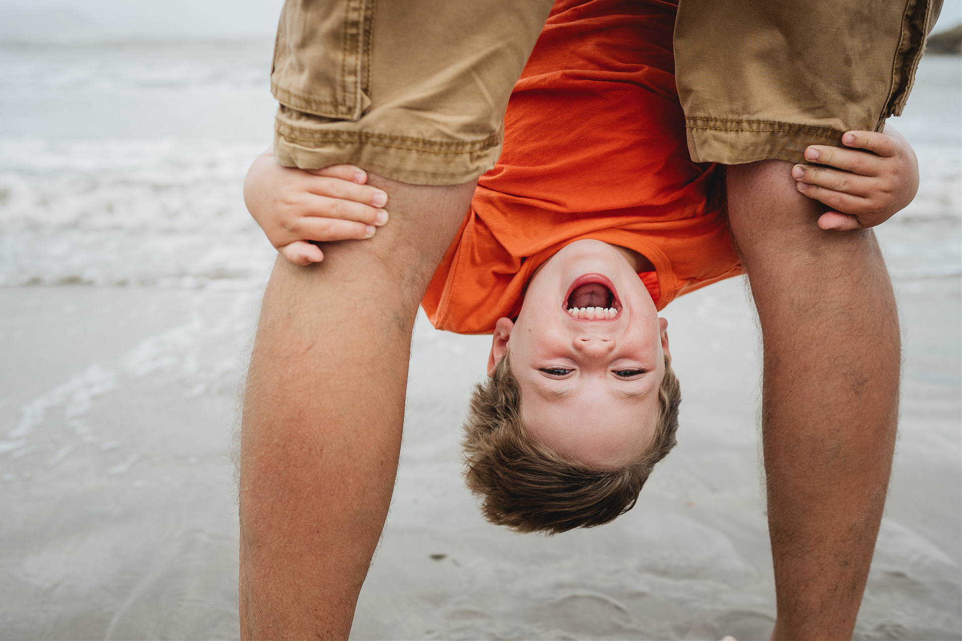 Devon family photography at the beach of a young boy laughing upside down between his father's legs