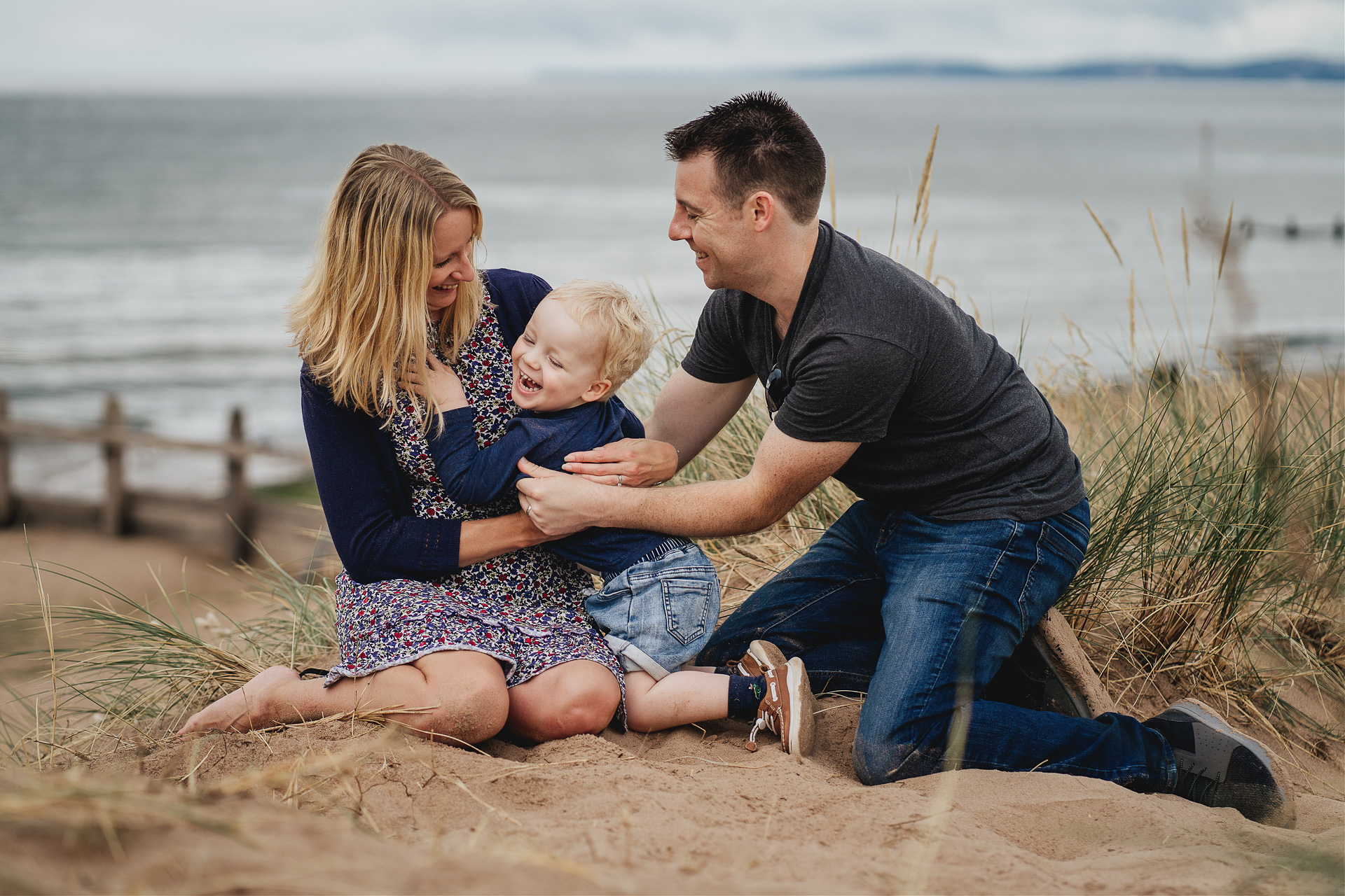 Devon family photography at the beach with two parents tickling their son and all laughing together on sand dunes at Dawlish Warren