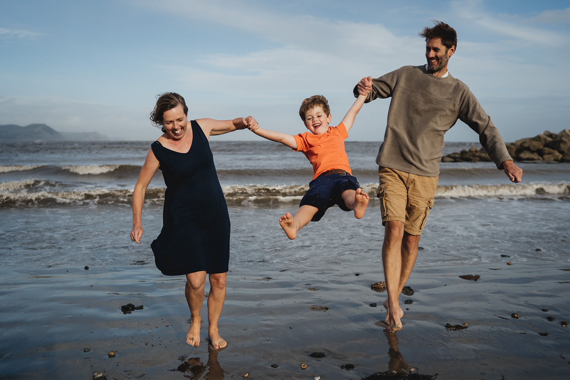 Best Devon family photography at the beach with parents swinging a young son into the air at the beach