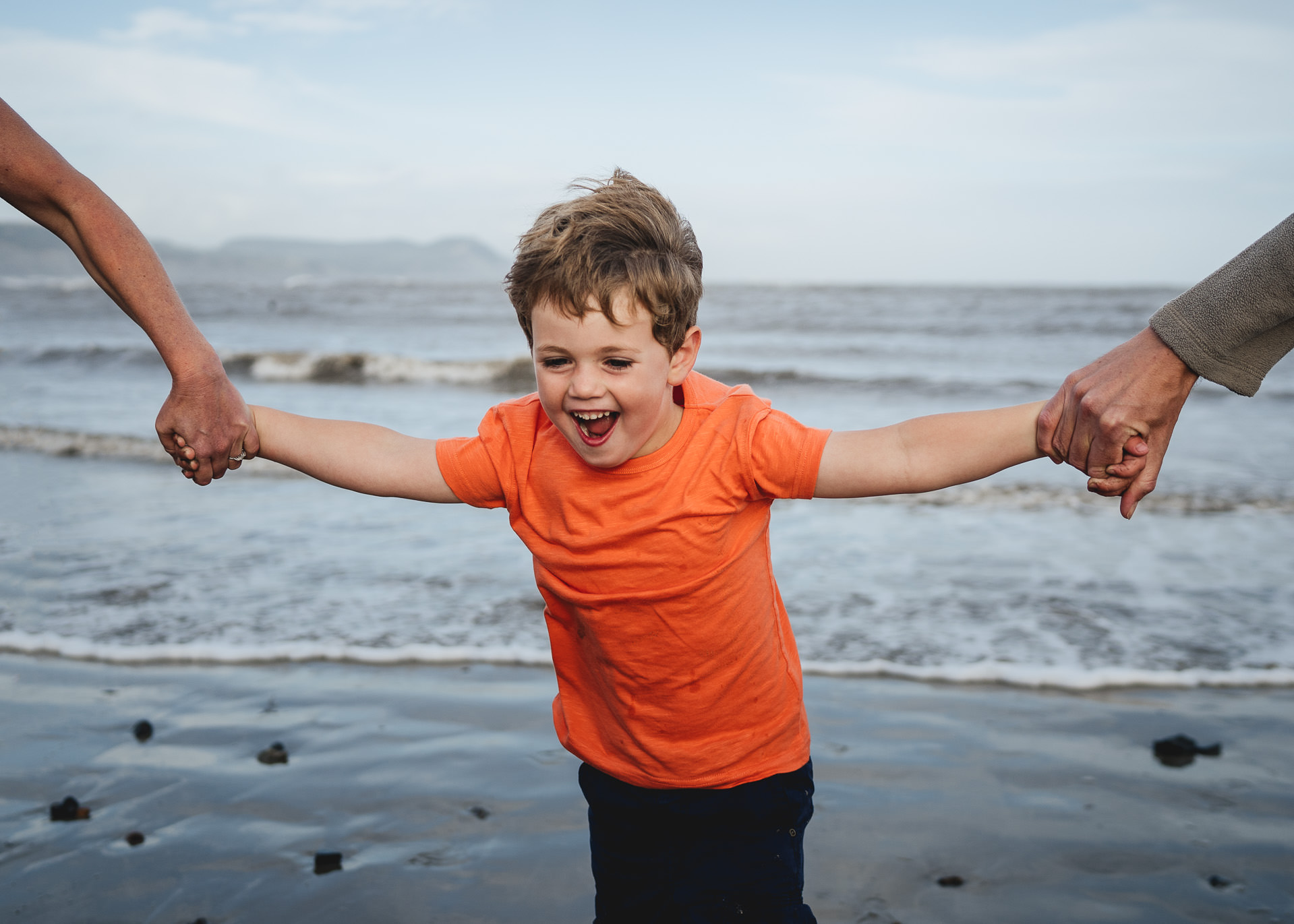 Devon family photography at the beach of a young boy laughing holding parents' hands