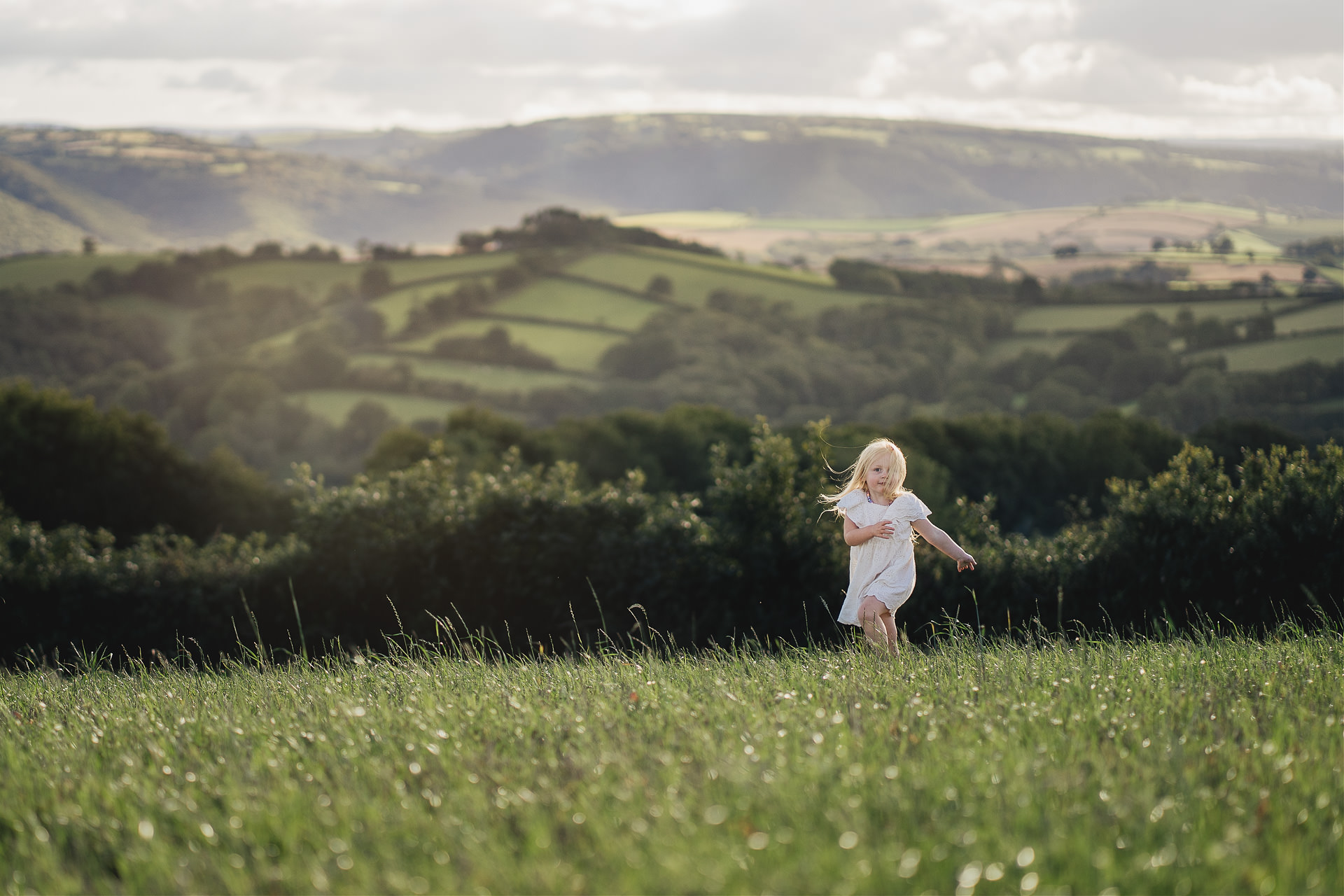 Best Devon family photography: a portrait of a young girl in a white dress running across a field with Devon hills behind