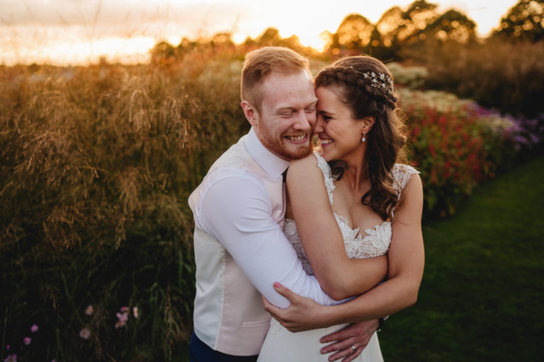 A bride and groom cuddling and smiling with a sunset and flowers behind them