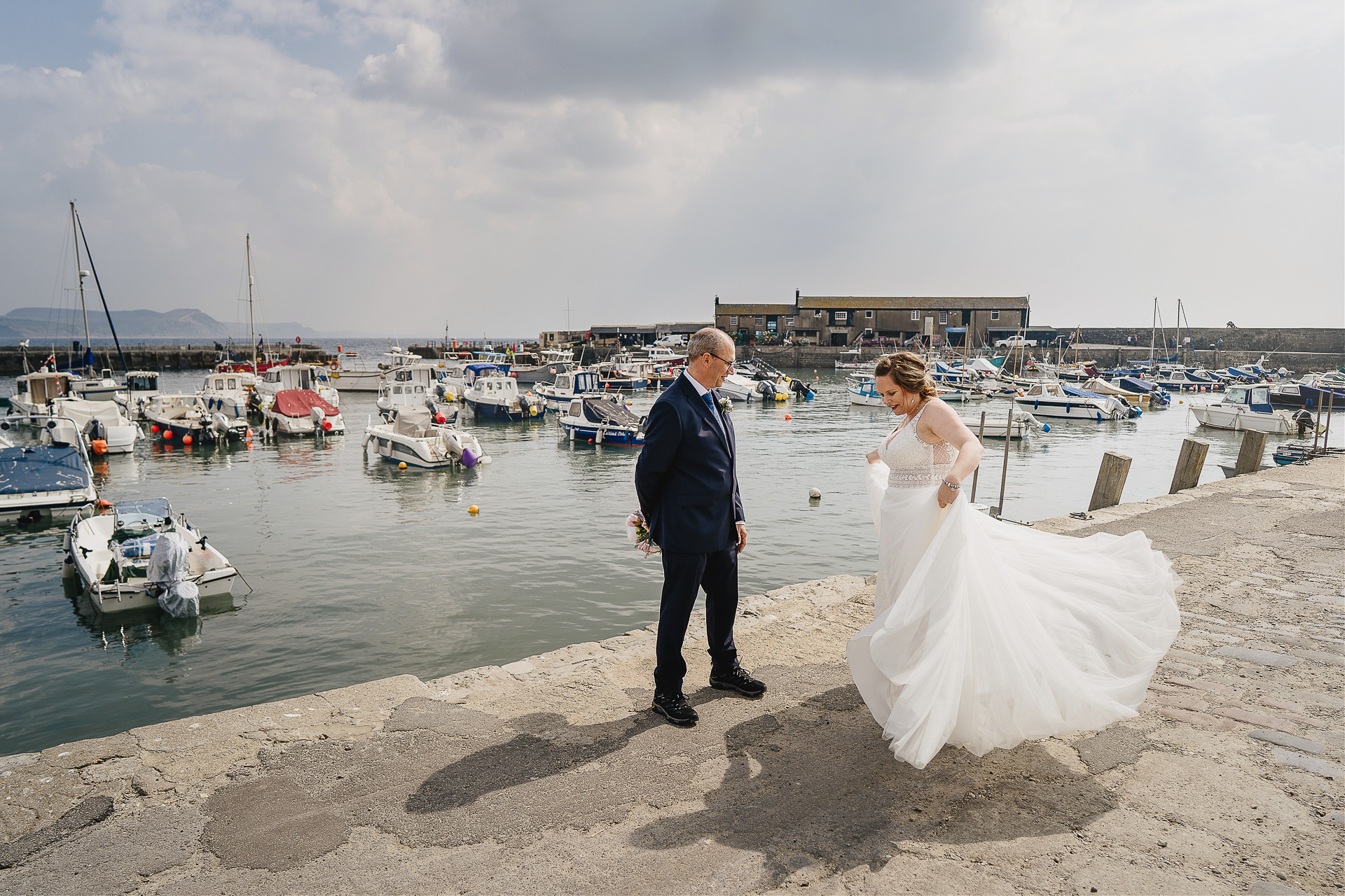 Groom watching bride twirling in her wedding dress by the harbour at Lyme Regis
