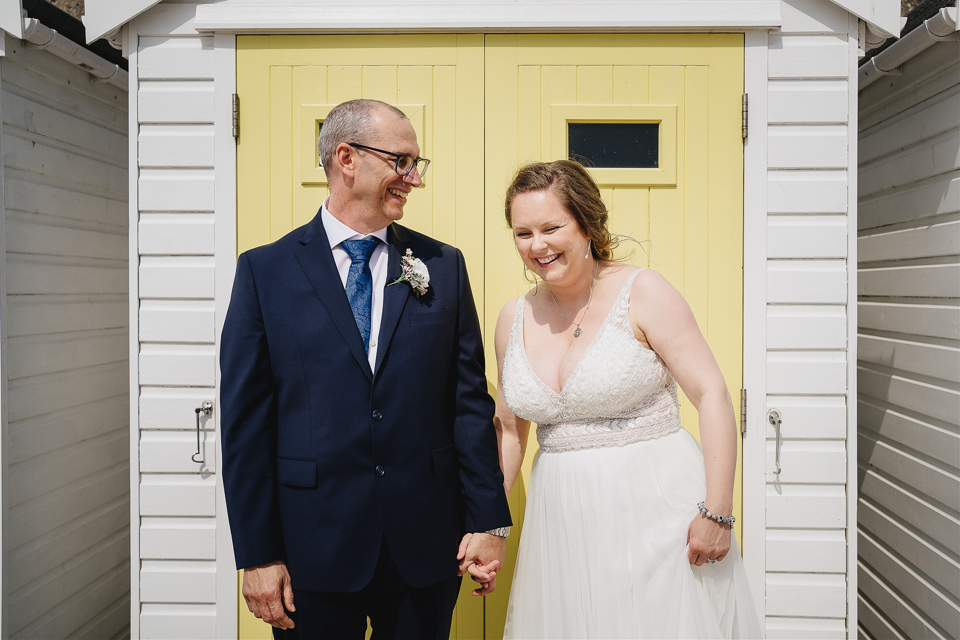 A bride and groom laughing together in front of a yellow beach hut
