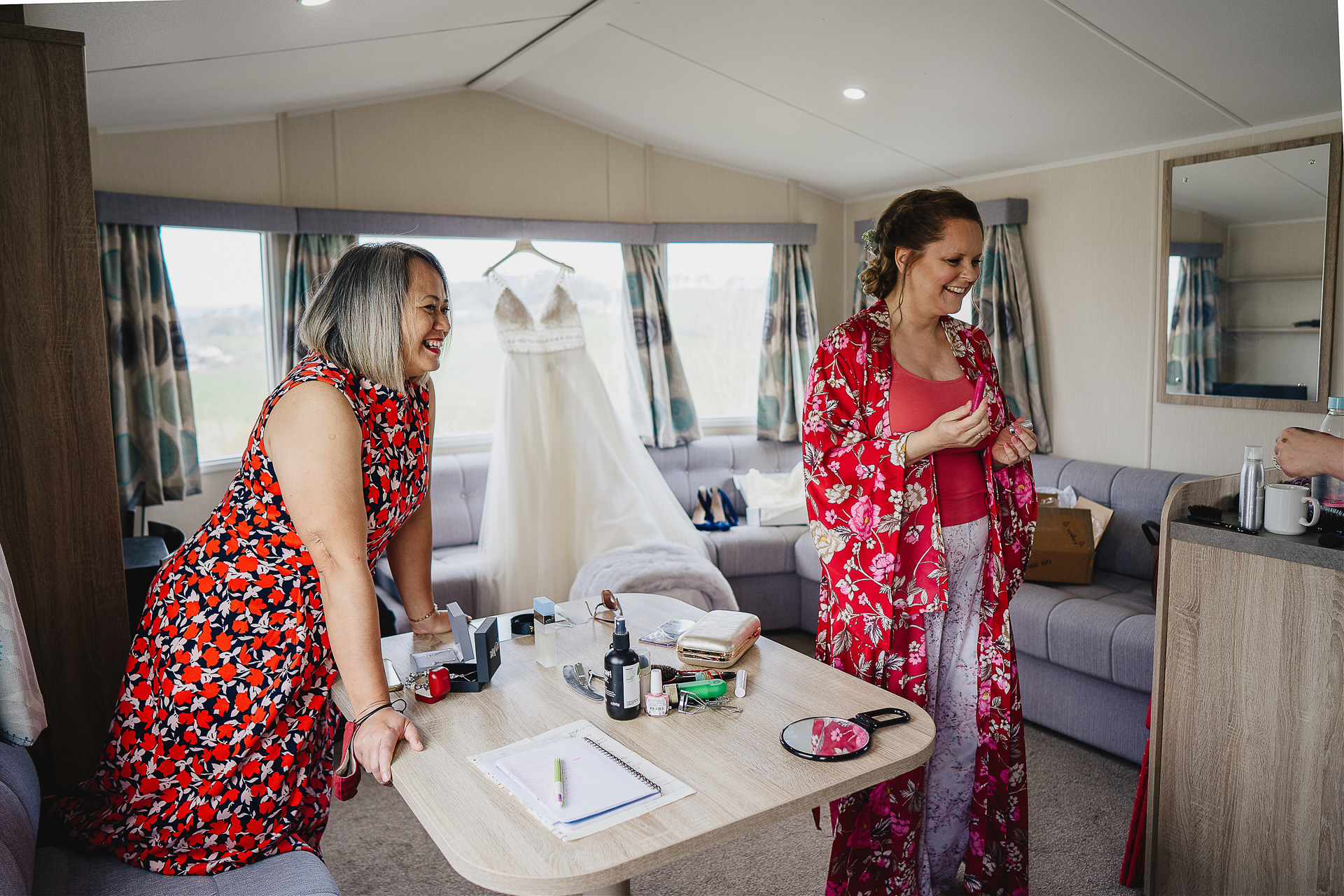 Bride and friend smiling in a caravan with wedding dress hanging behind them