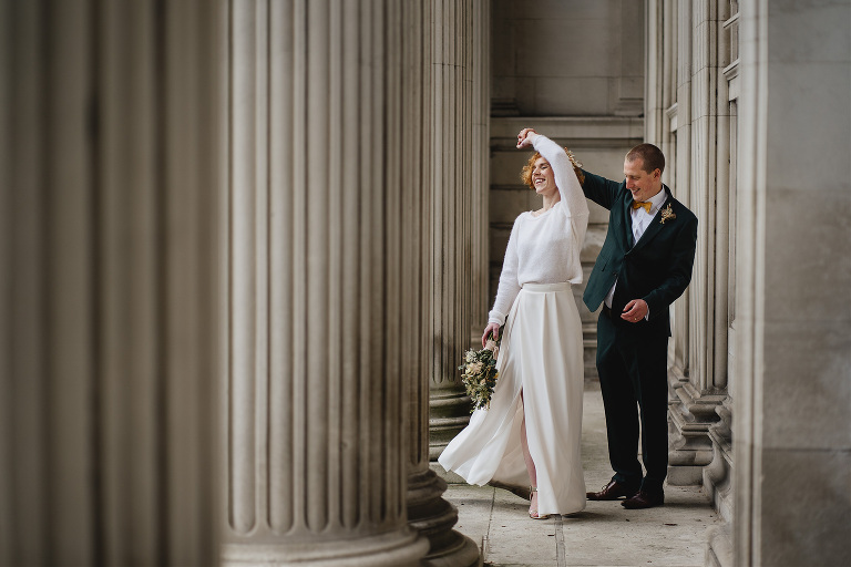 A groom in a green suit, twirling a bride around amongst some white pillars. 