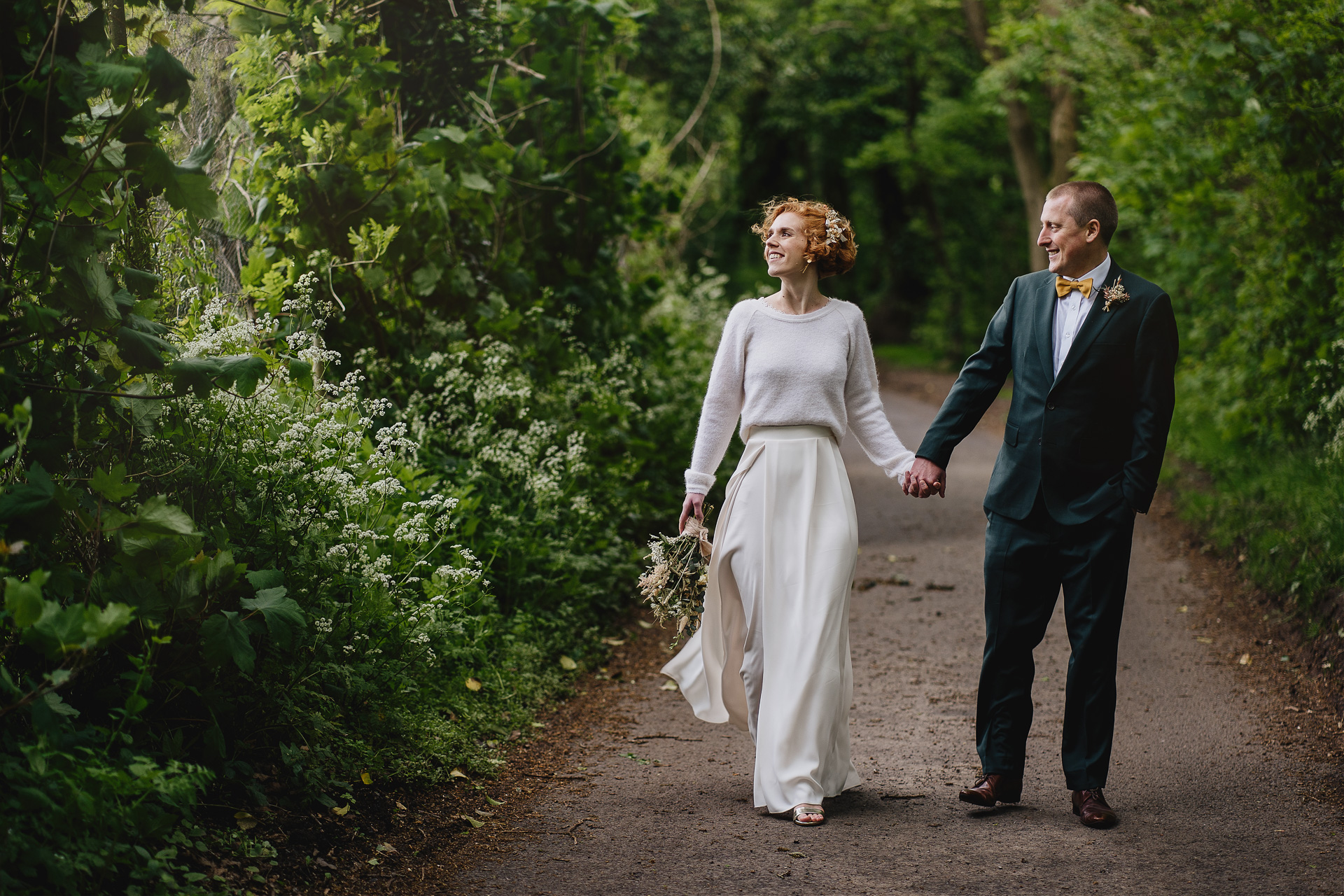 A bride and groom walking together intimately, hand in hand, along a country lane with trees all around. 
