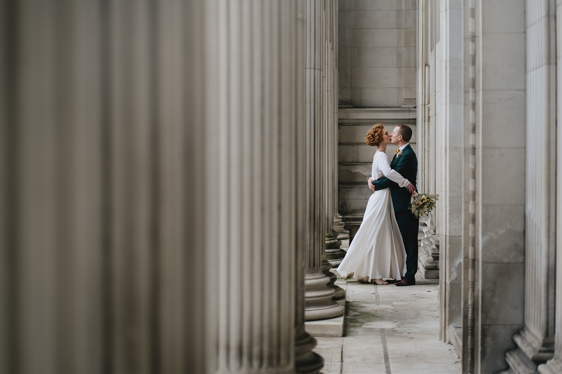 An intimate wedding celebration, with a bride and groom kissing amongst some white pillars, with nobody else around. 