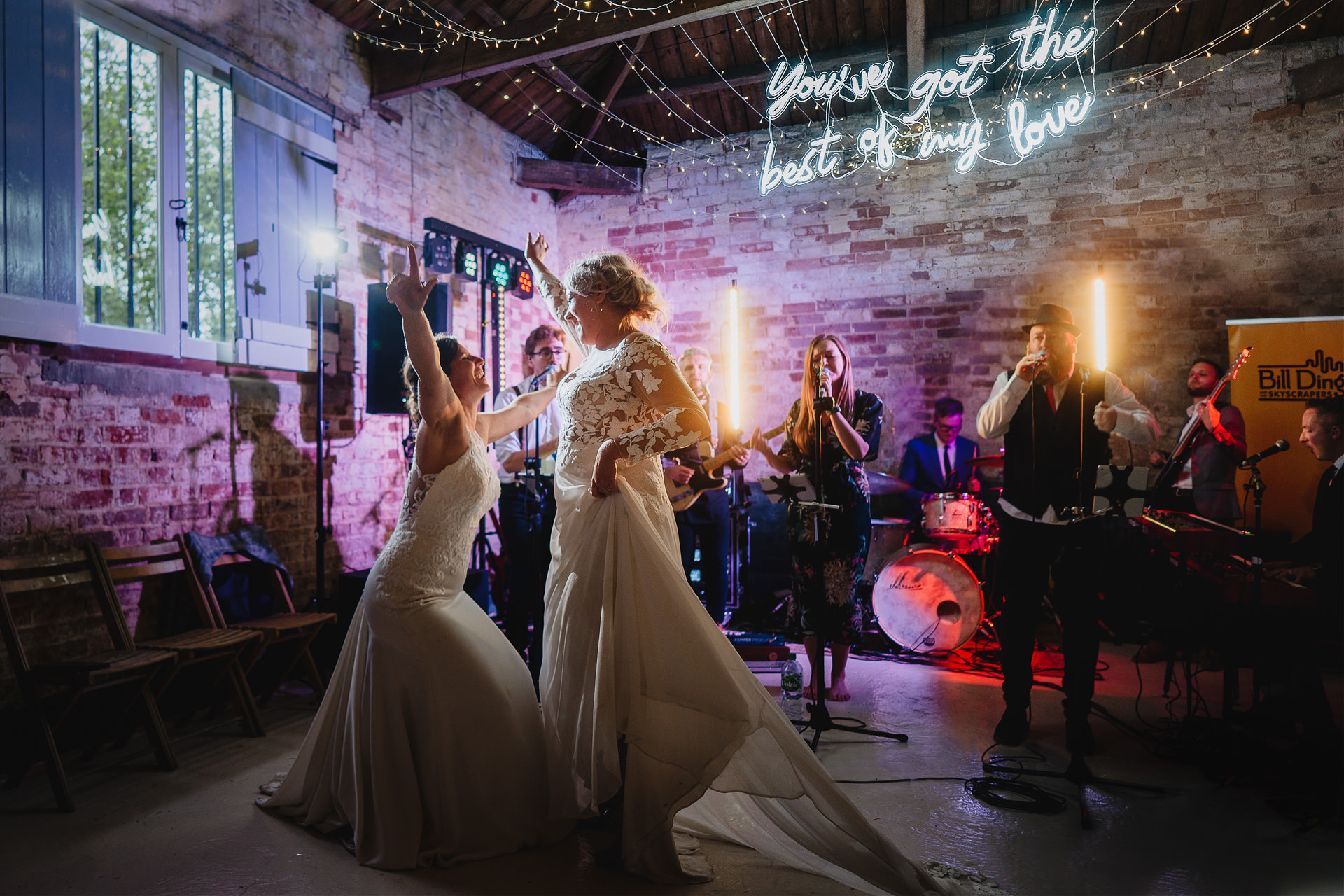 Two brides on the dance floor at their Cadhay wedding with the band behind them. 