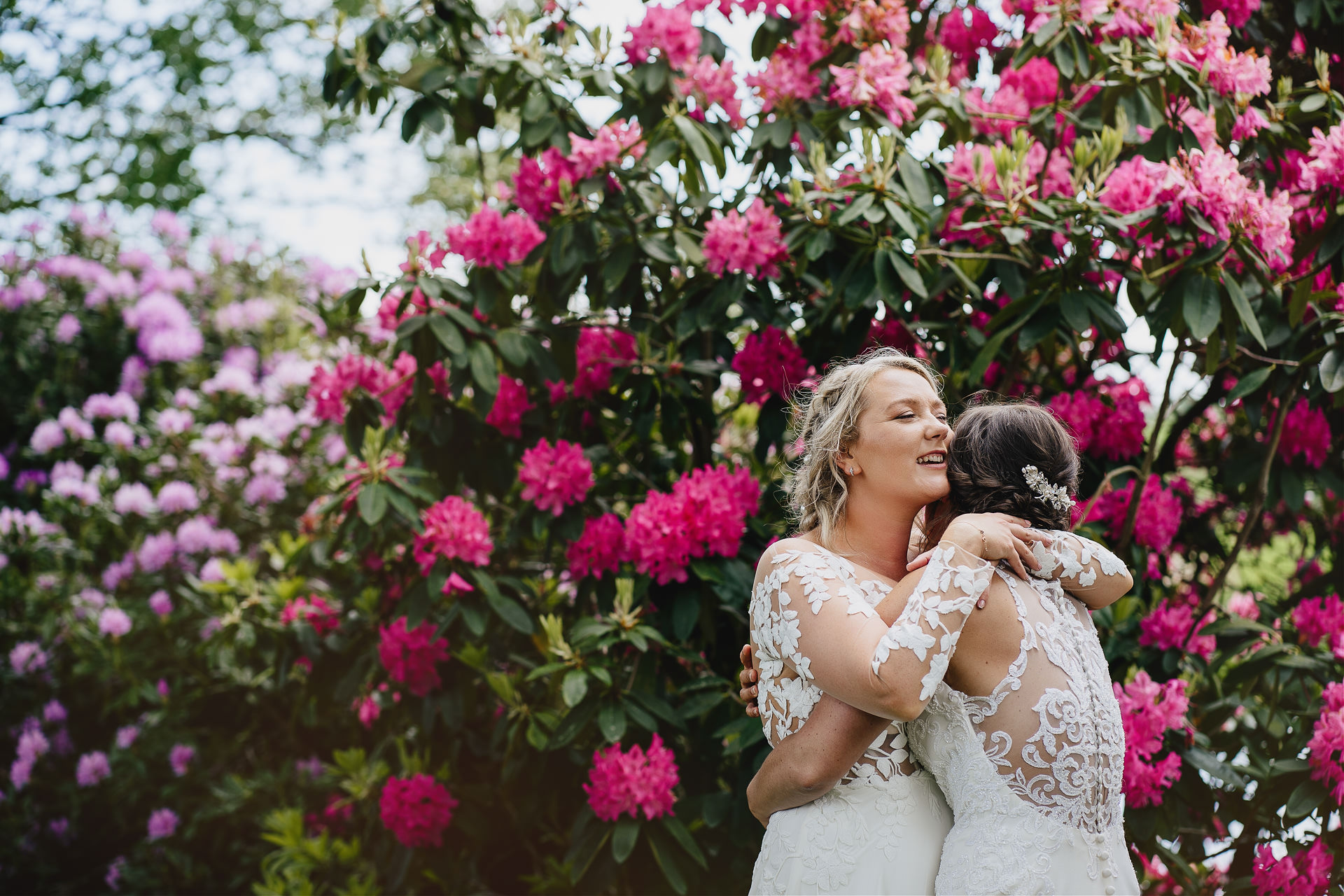 Two brides cuddling together in front of a beautiful display of large pink flowers at their Cadhay wedding in Devon