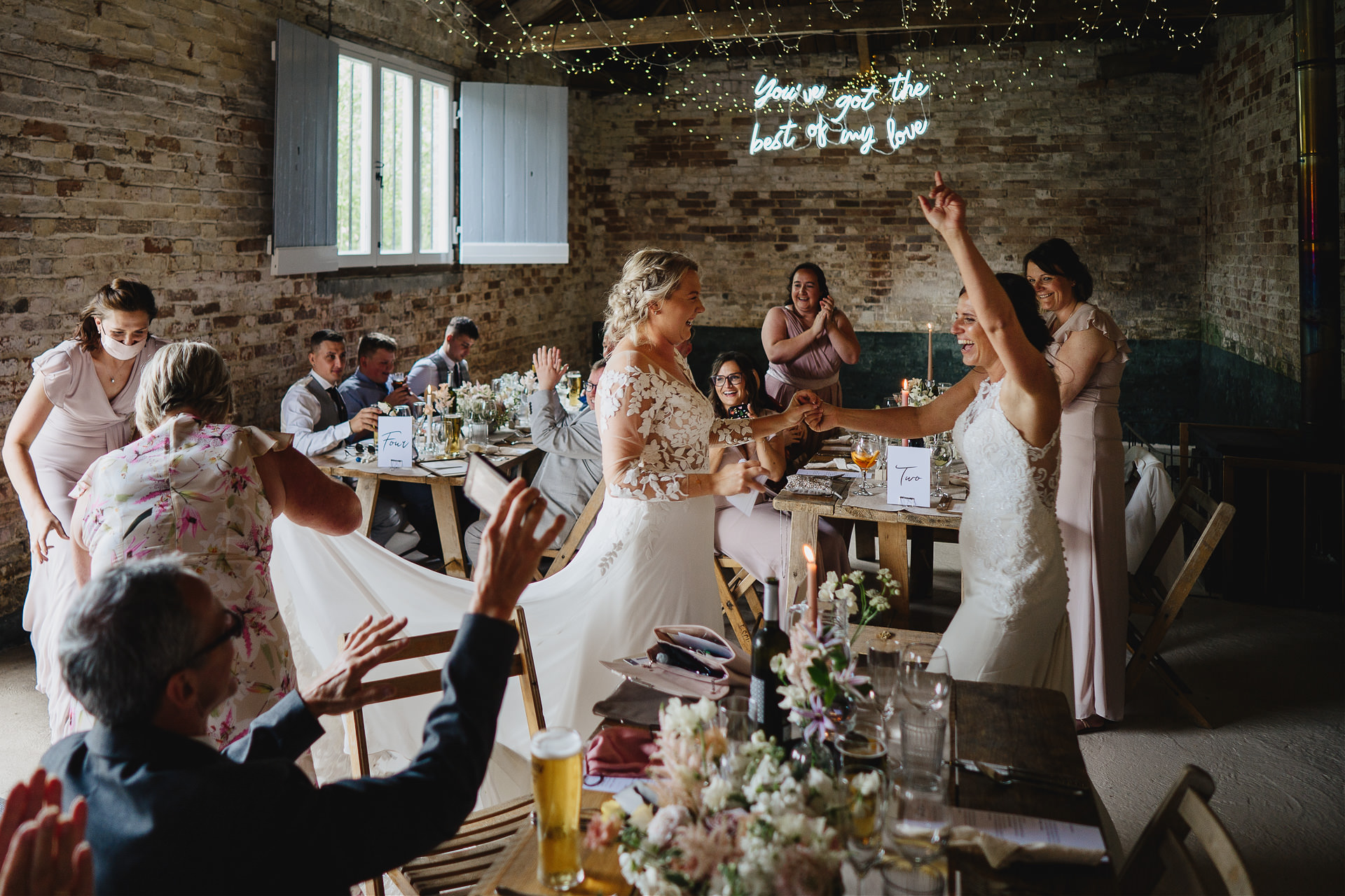 Two brides entering their wedding breakfast with guests cheering and clapping in a rustic barn