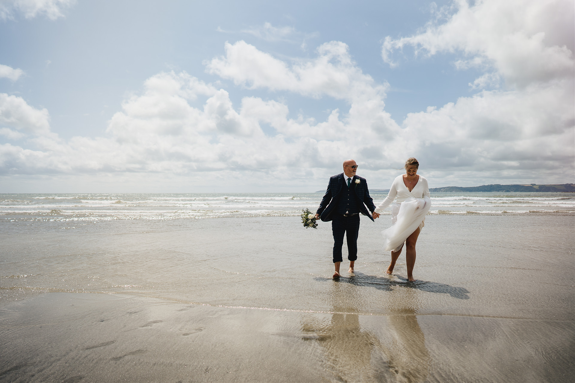 A bride and groom in wedding clothes, walking barefoot in the sea in Cornwall