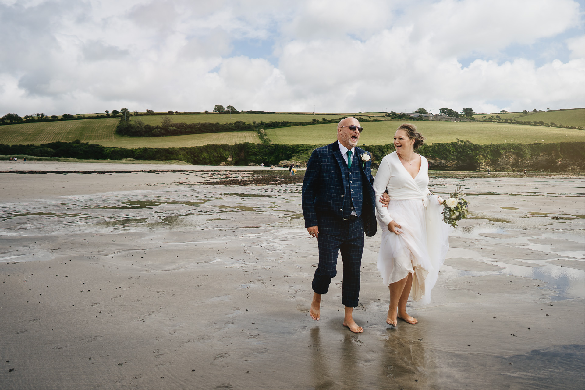 A bride and groom walking and laughing on a Cornish beach
