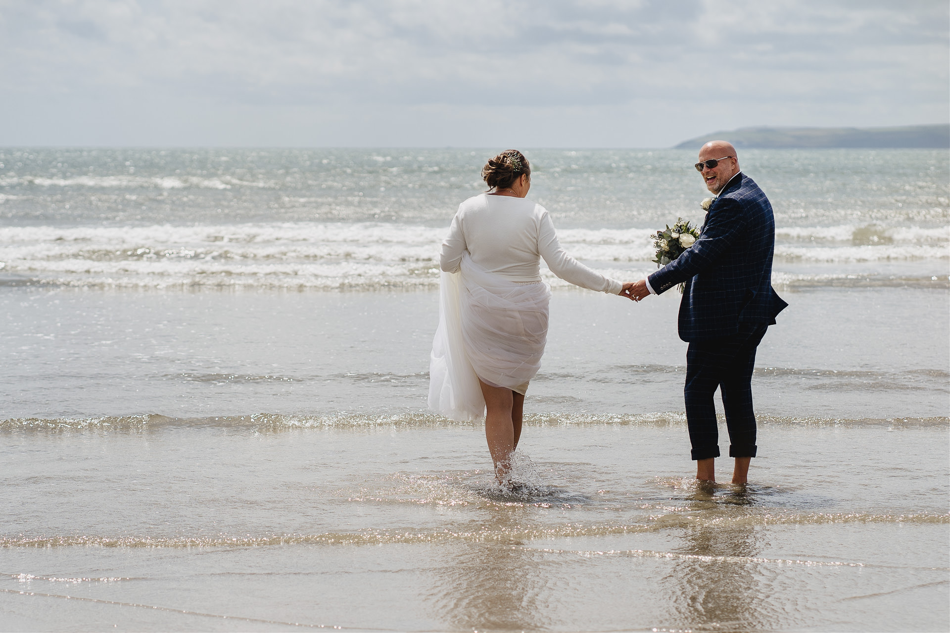 A bride and groom in wedding clothes, paddling barefoot in the sea in Cornwall