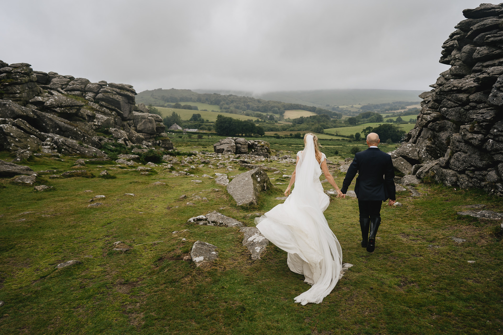Bride and groom walking hand in hand across Dartmoor with views across the hills and rocks