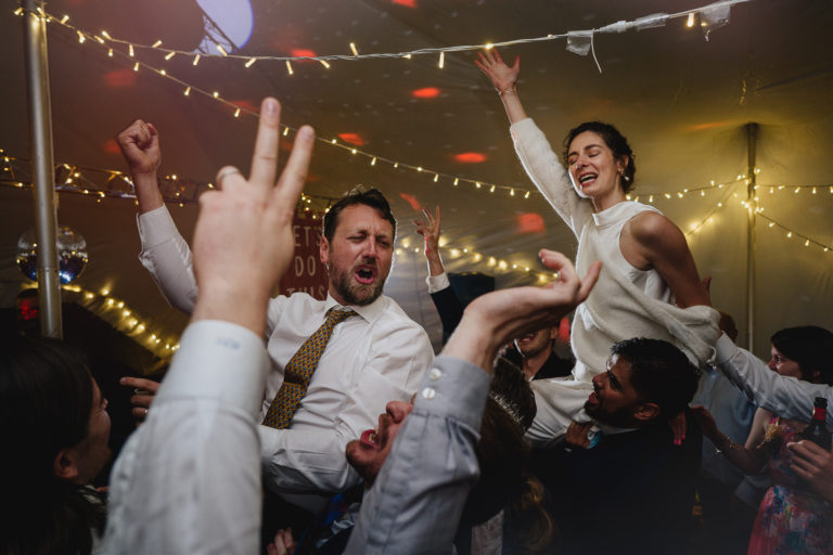 Bride and groom on a packed dance floor in a marquee, lifted up on the shoulders of weddings guests