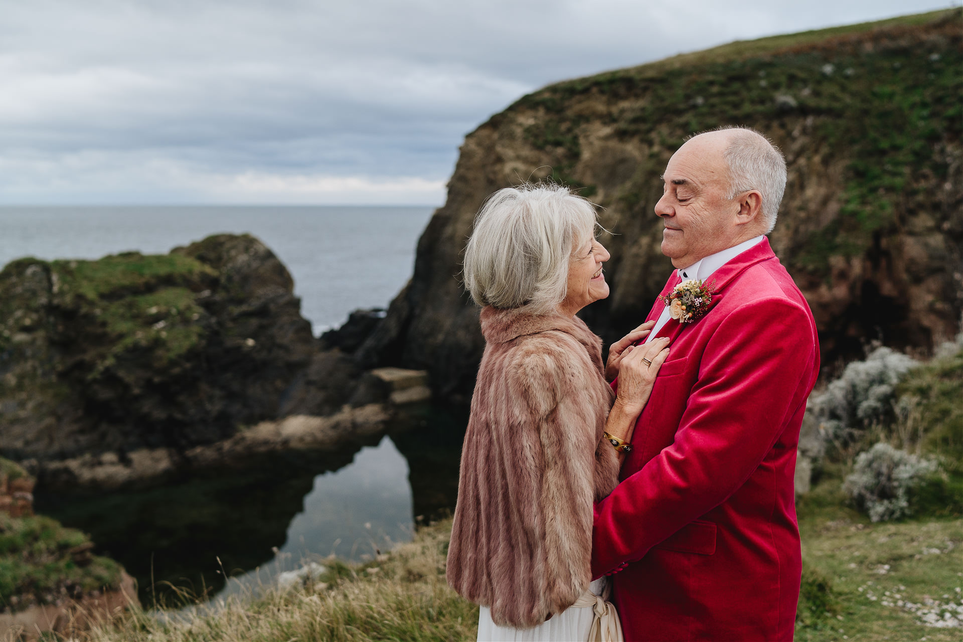 A bride with a fur cape and a groom in a pink velvet jacket by the mermaid pool at Burgh Island, smiling at each other before their elopement wedding