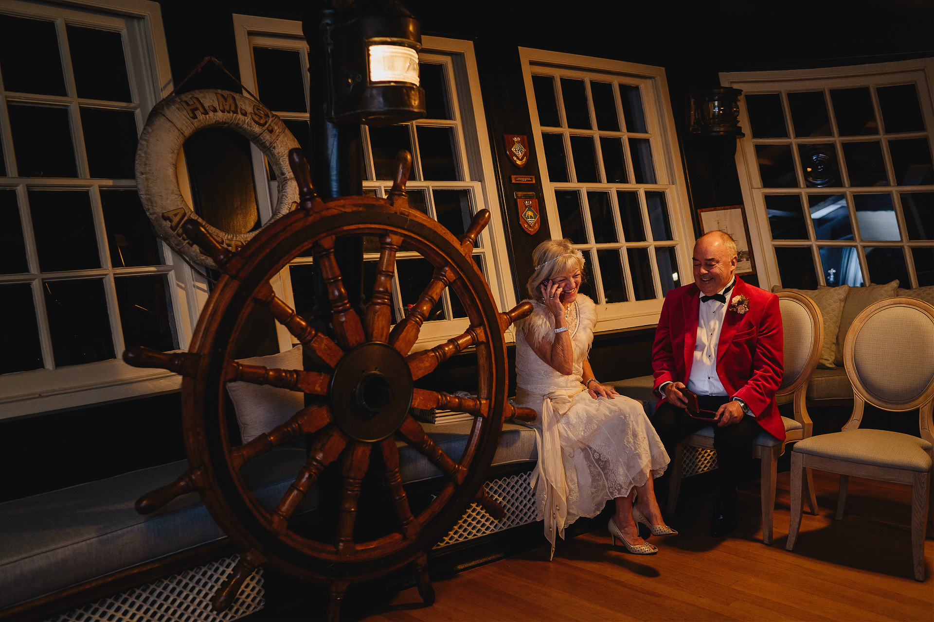 A bride and groom laughing together and talking on the phone next to a large ships wheel at Burgh Island