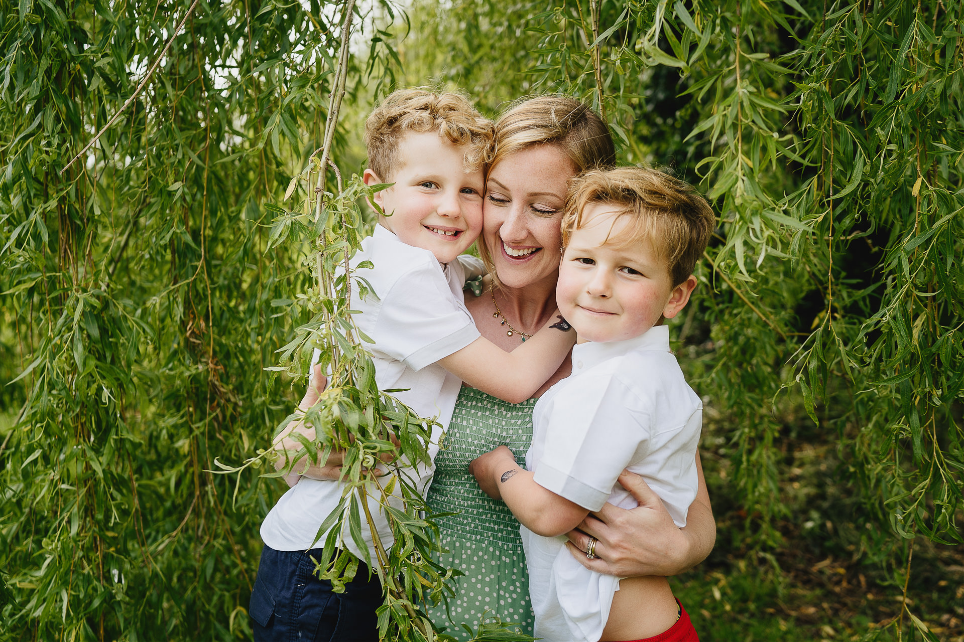 Two boys cuddling their mother amongst willow tree leaves