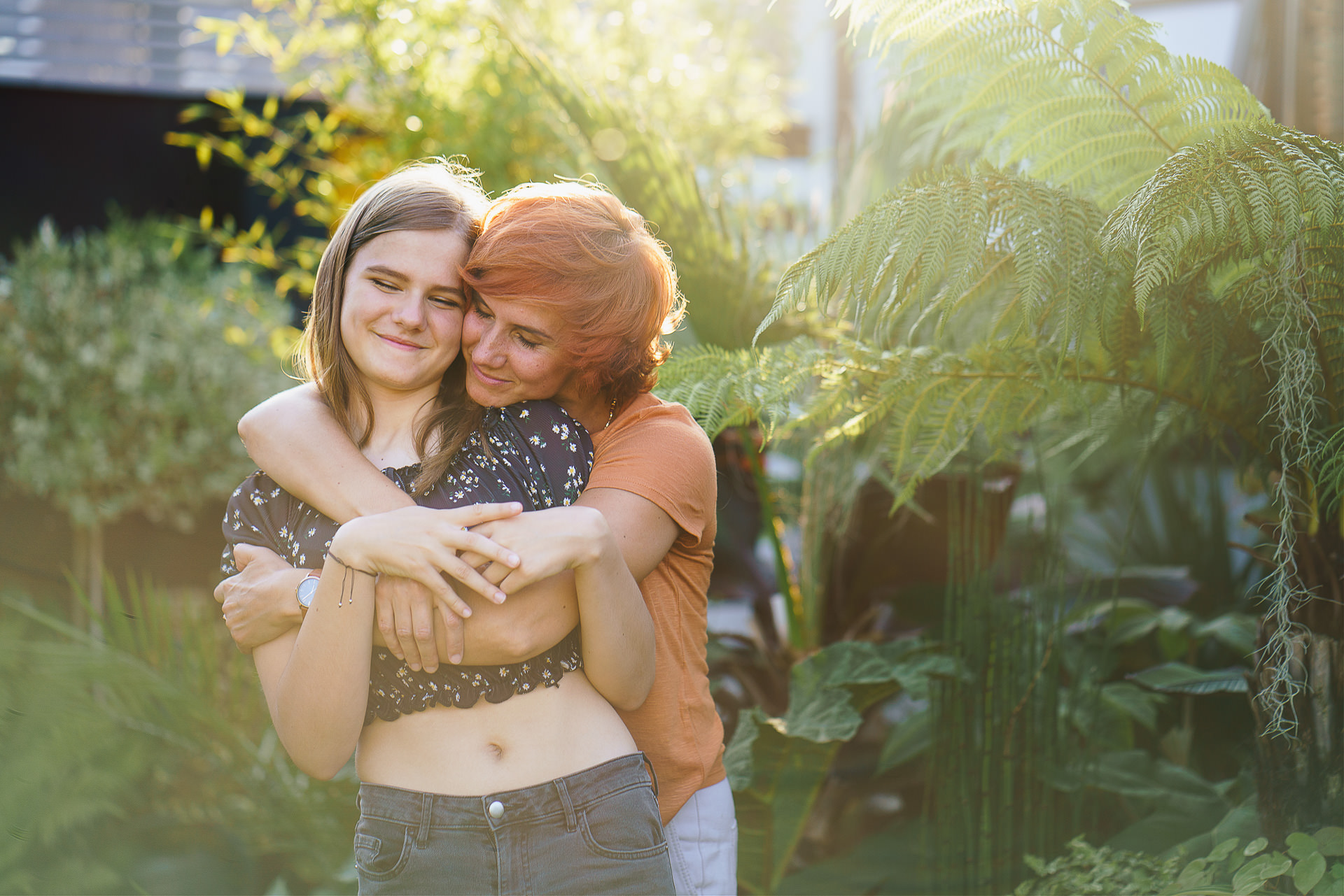 A mother cuddling her teenage daughter in front of ferns and greenery