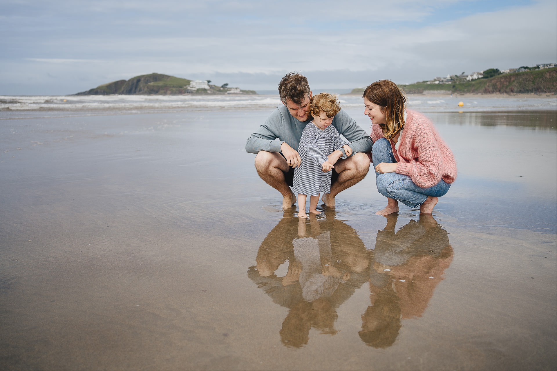 A family photography session on a beautiful Devon beach with two parents and a young child squatting in the sand